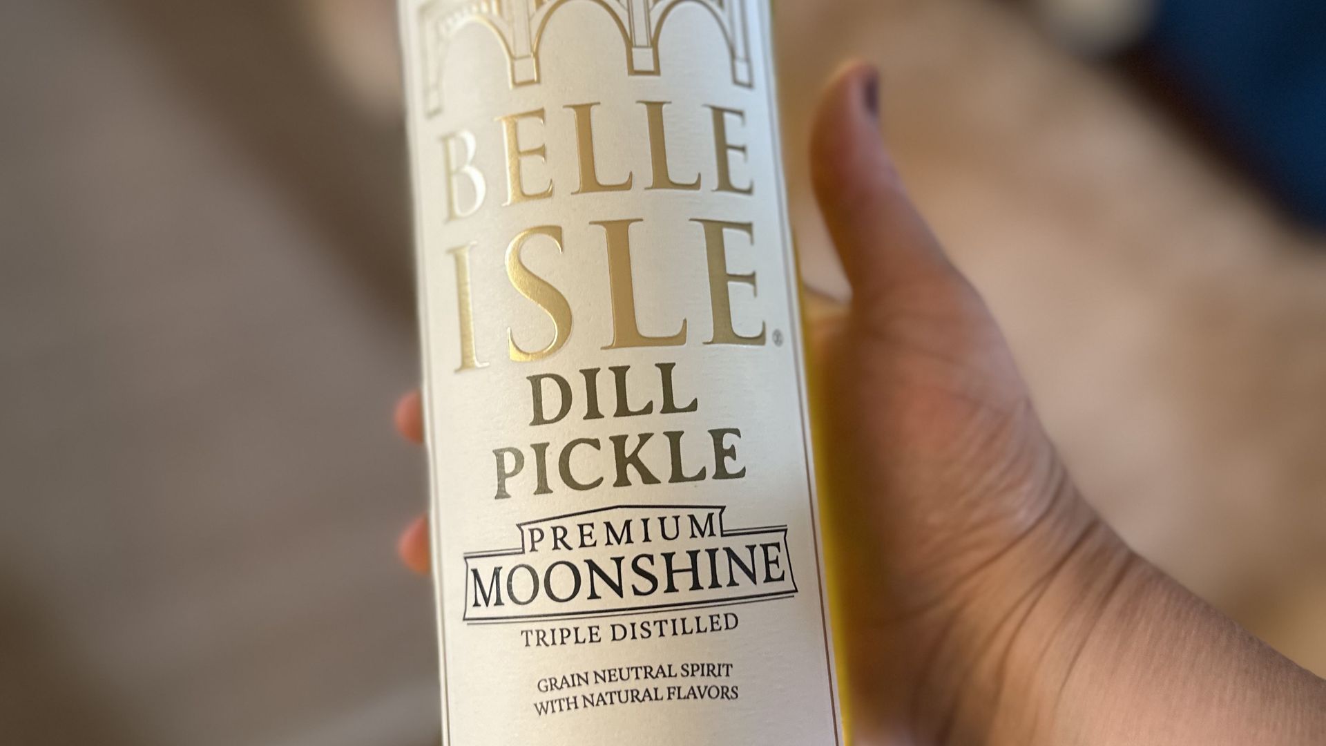 Hand holding a tall bottle of Belle Isle Dill Pickle Moonshine. White label with gold embossed text reading "BELLE ISLE" and "DILL PICKLE"; blurred indoor background.