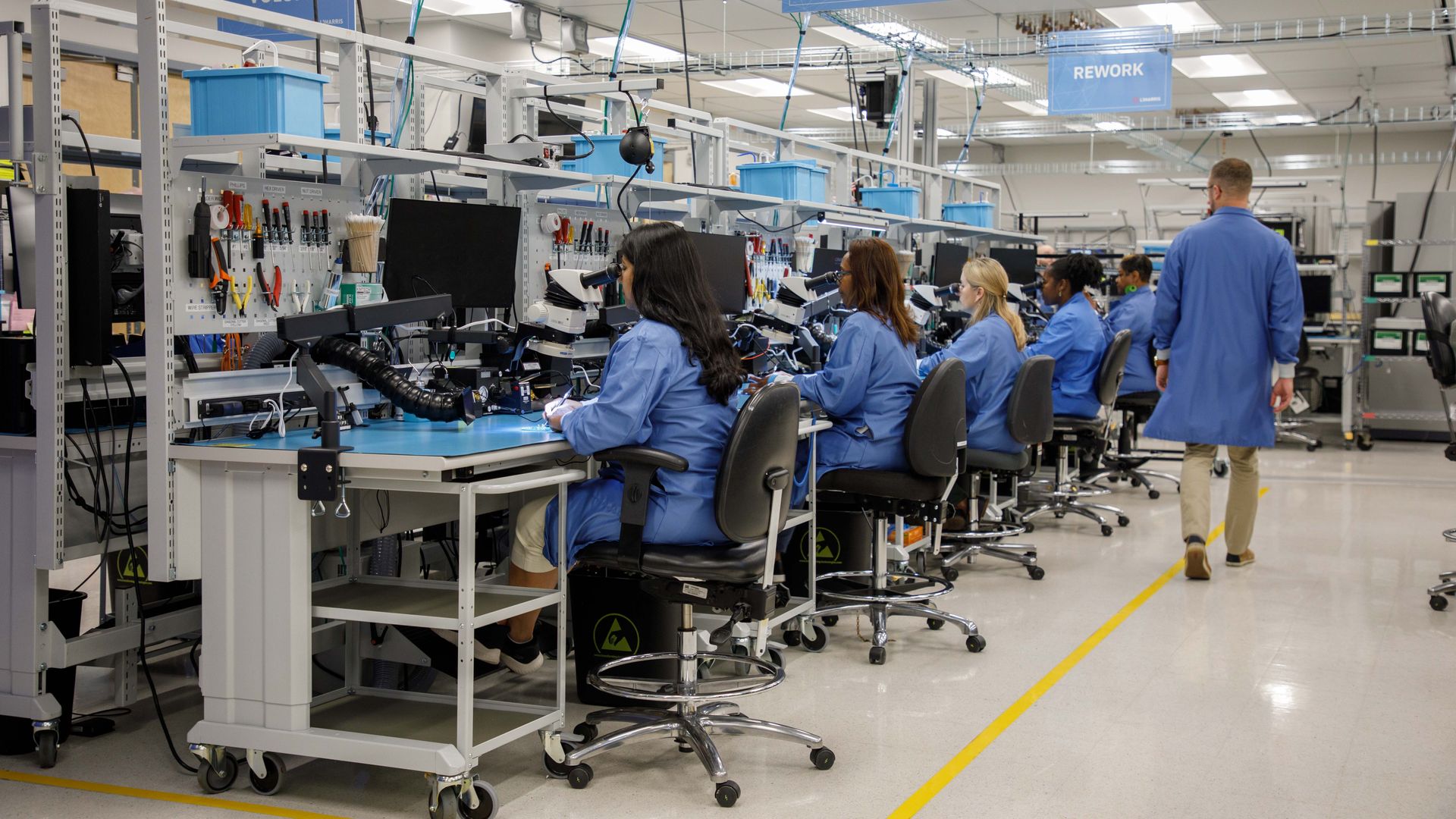 People are seated at workbenches. They wear blue overcoats. They're in a bright lab. There's a yellow  line, as well.