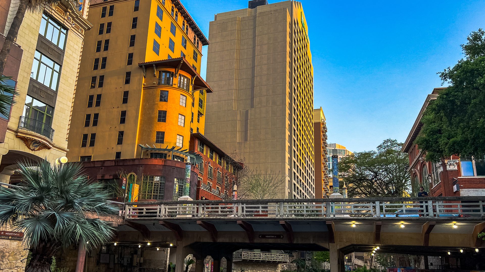 View of San Antonio's River Walk at dusk, with a footbridge crossing over the river, surrounded by colorful historic and modern buildings, palm trees, and clear blue skies.