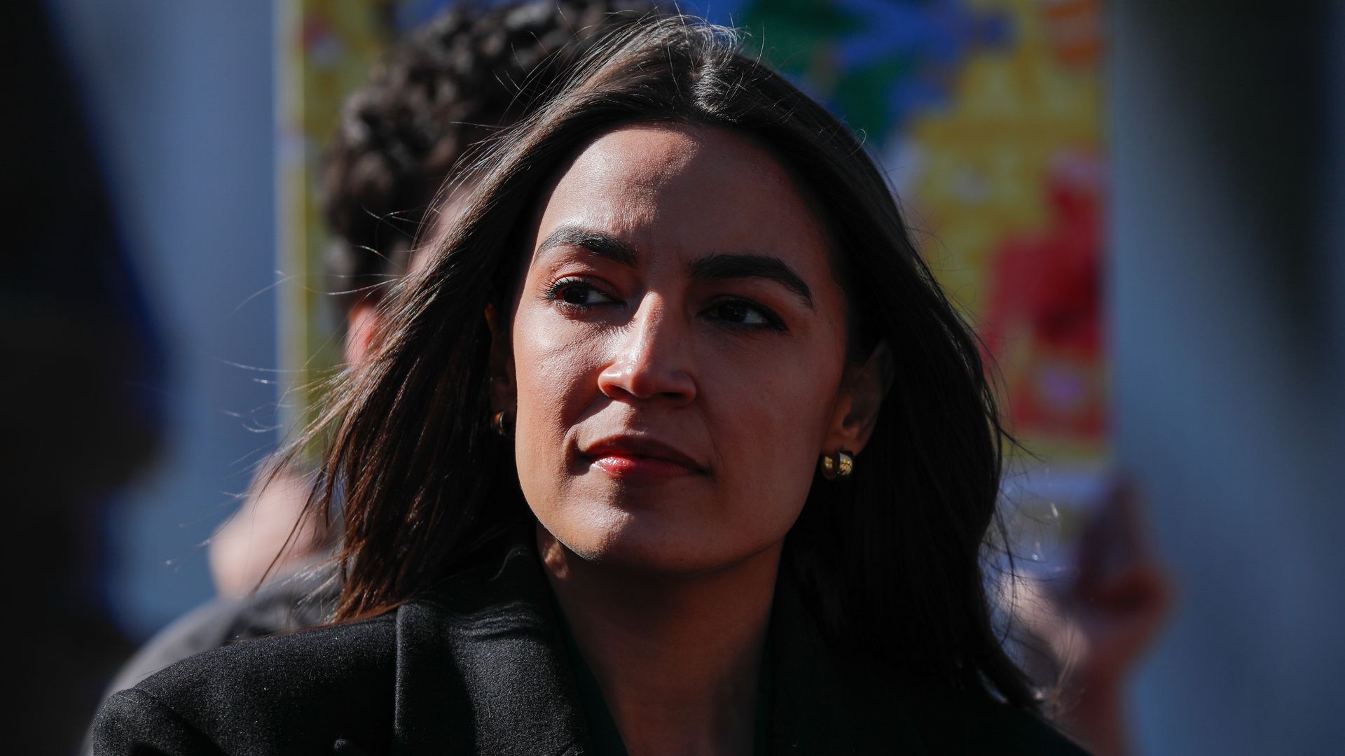 Rep. Alexandria Ocasio Cortez, wearing a black coat and standing in front of a multi-colored poster.