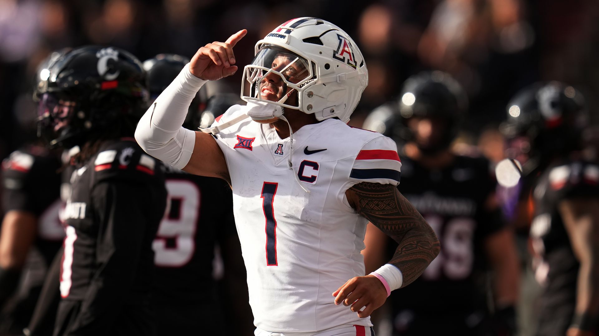 A football player in a white uniform with a blue "1" on the front and a red and blue "A" on the side of the helmet holds up his right hand and points his index finger up in a victorious gesture. Opposing players in black uniforms are in the background.