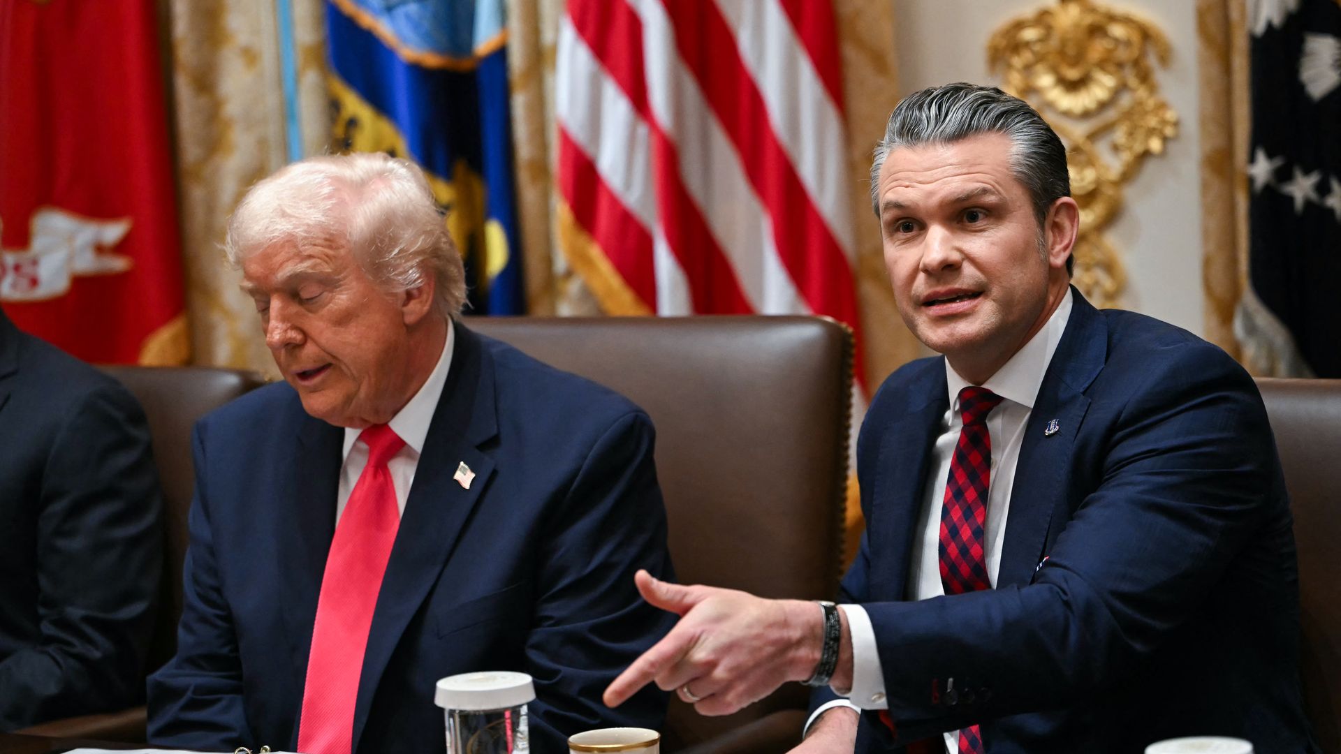 Pete Hegseth, wearing a dark suit and a tie, points as he speaks, sitting at a table next to Donald Trump, who is wearing a dark suit and a red tie.