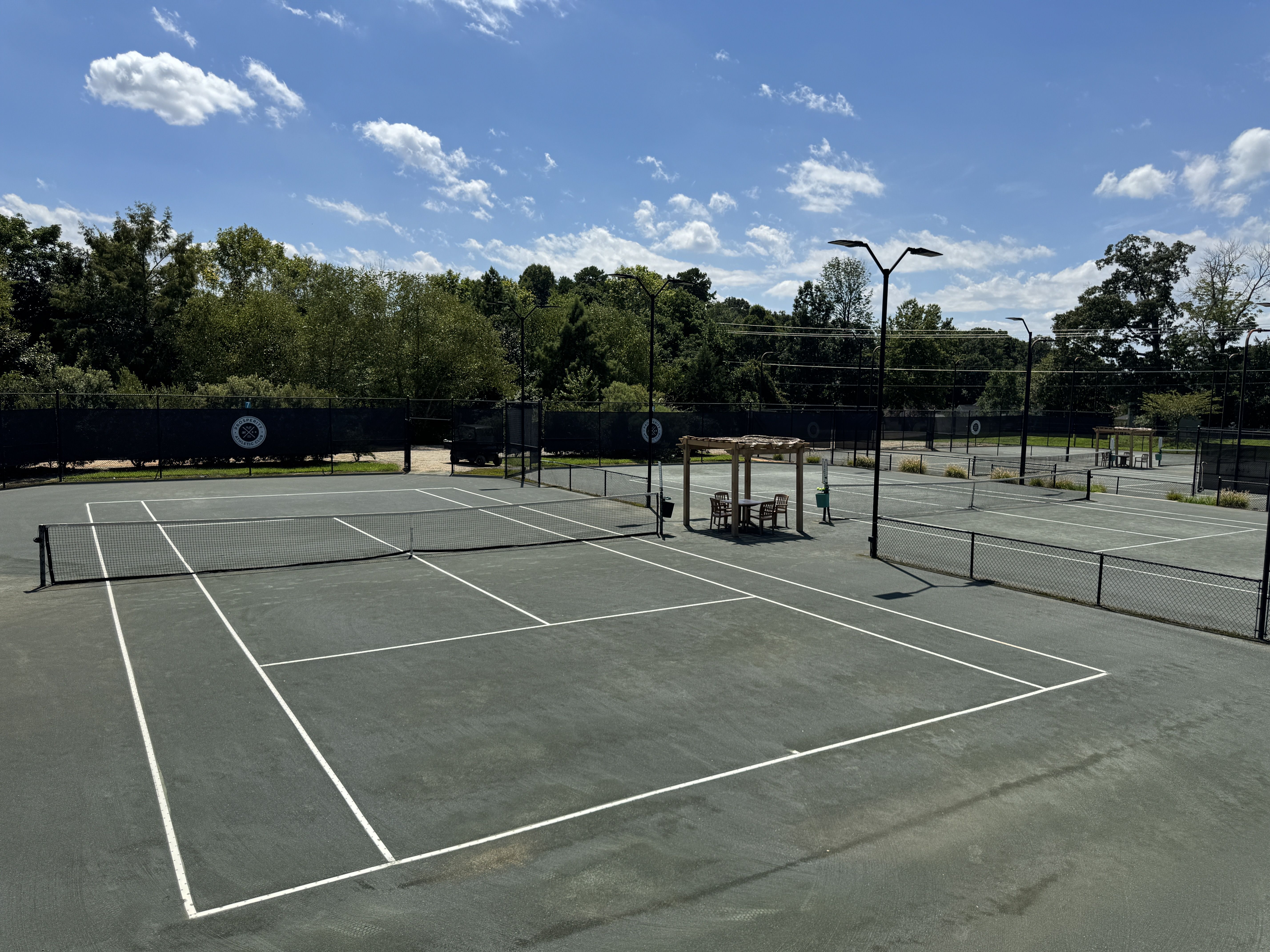 Empty outdoor tennis courts with grayish-green surfaces, white lines, nets, and wooden seating areas under small pergolas. Trees and blue sky with clouds in the background.