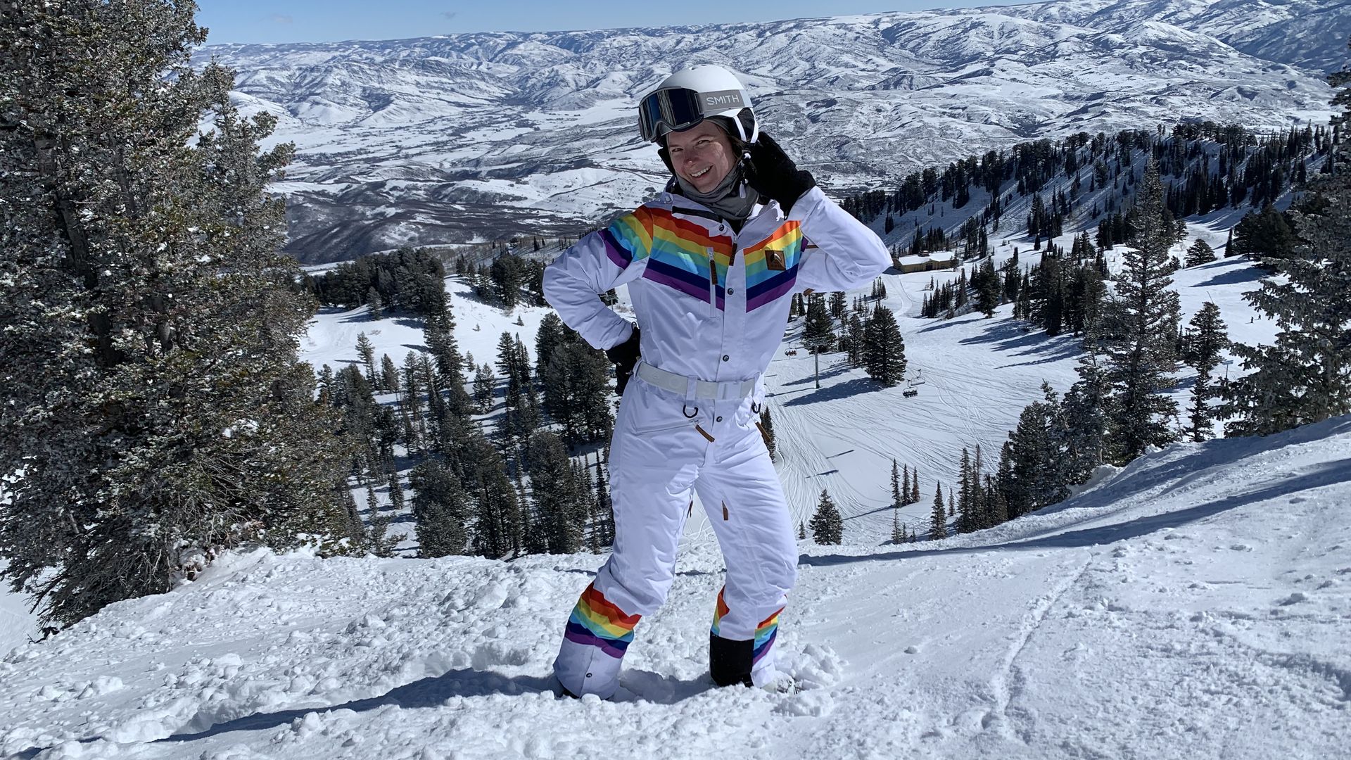 A photo of a smiling and posing woman wearing a white skiuit with rainbow colors on the shoulders and legs while standing on a ski slope