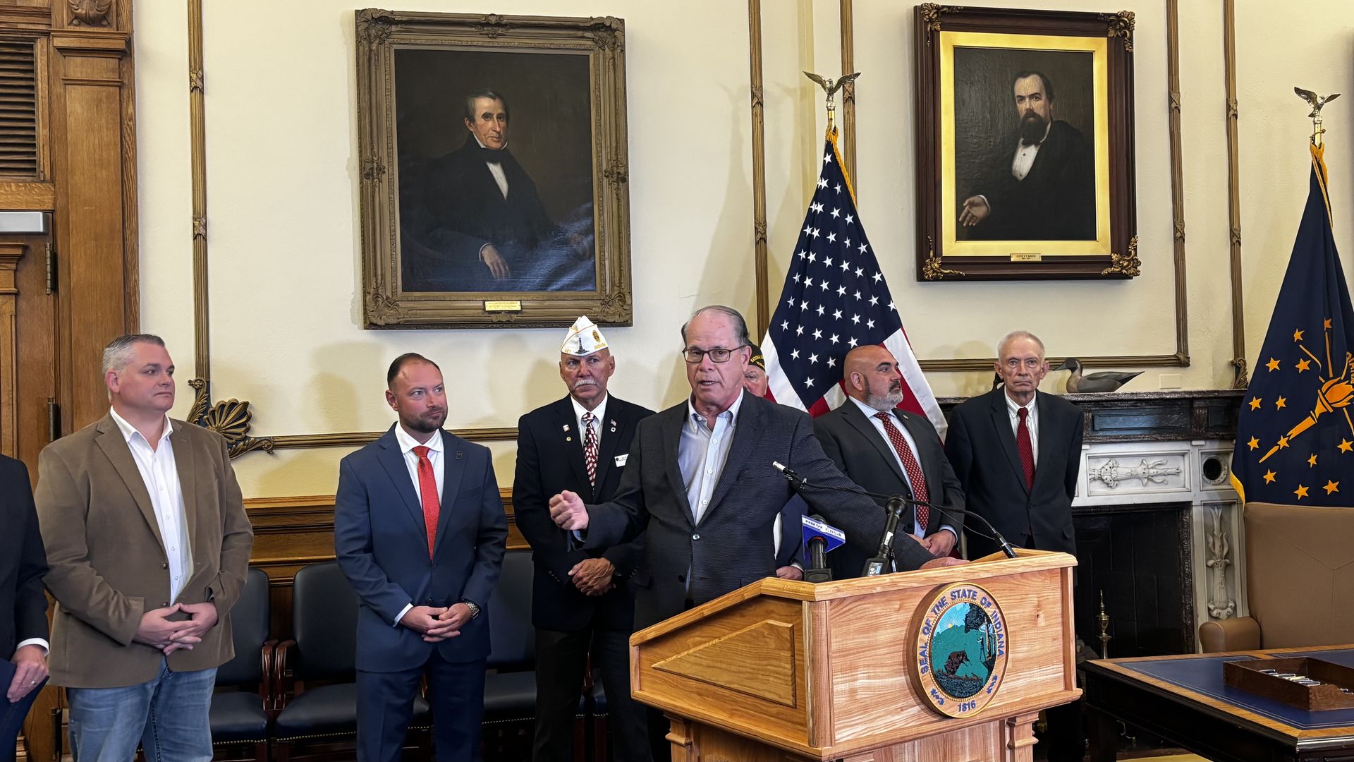 Formal press conference in a wood-paneled room; five men in suits stand by a wooden podium with a state seal. Behind them are two large gold-framed portraits and flags.