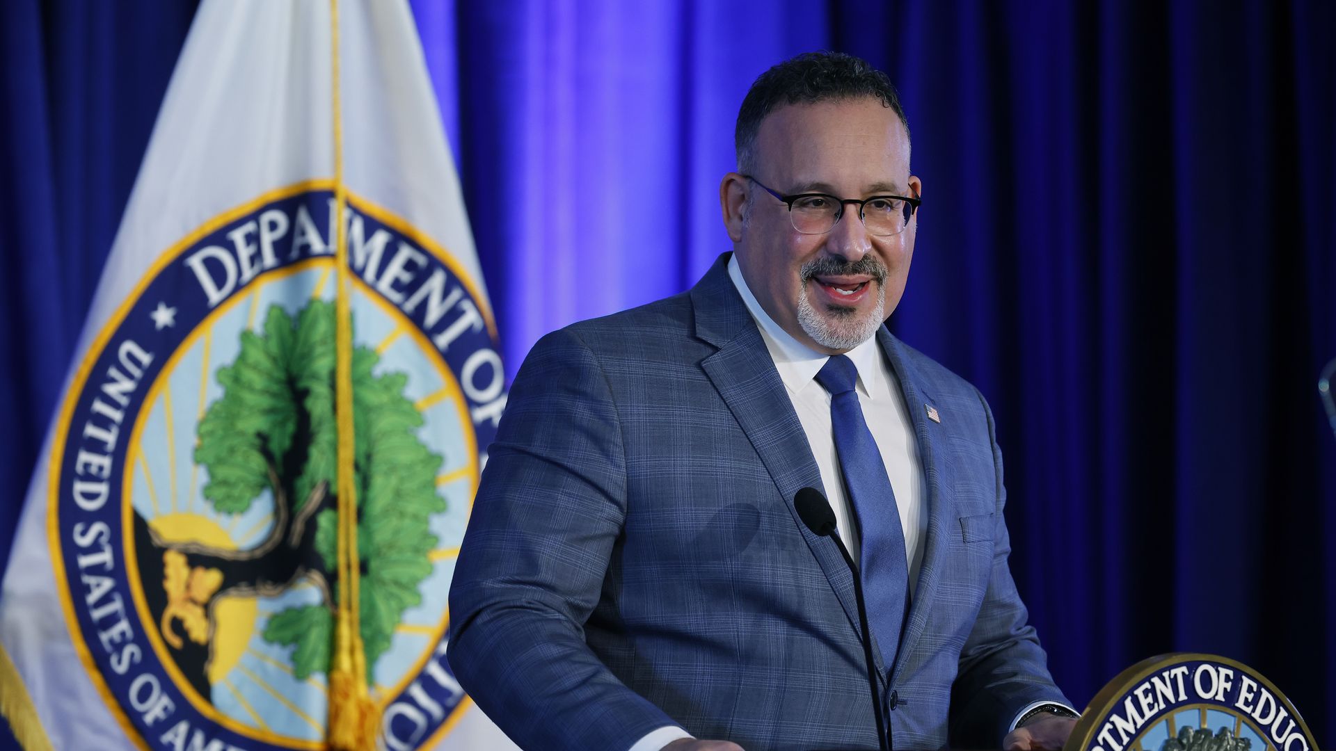 Photo of Miguel Cardona speaking from a podium as a flag with the Education Department seal in the back
