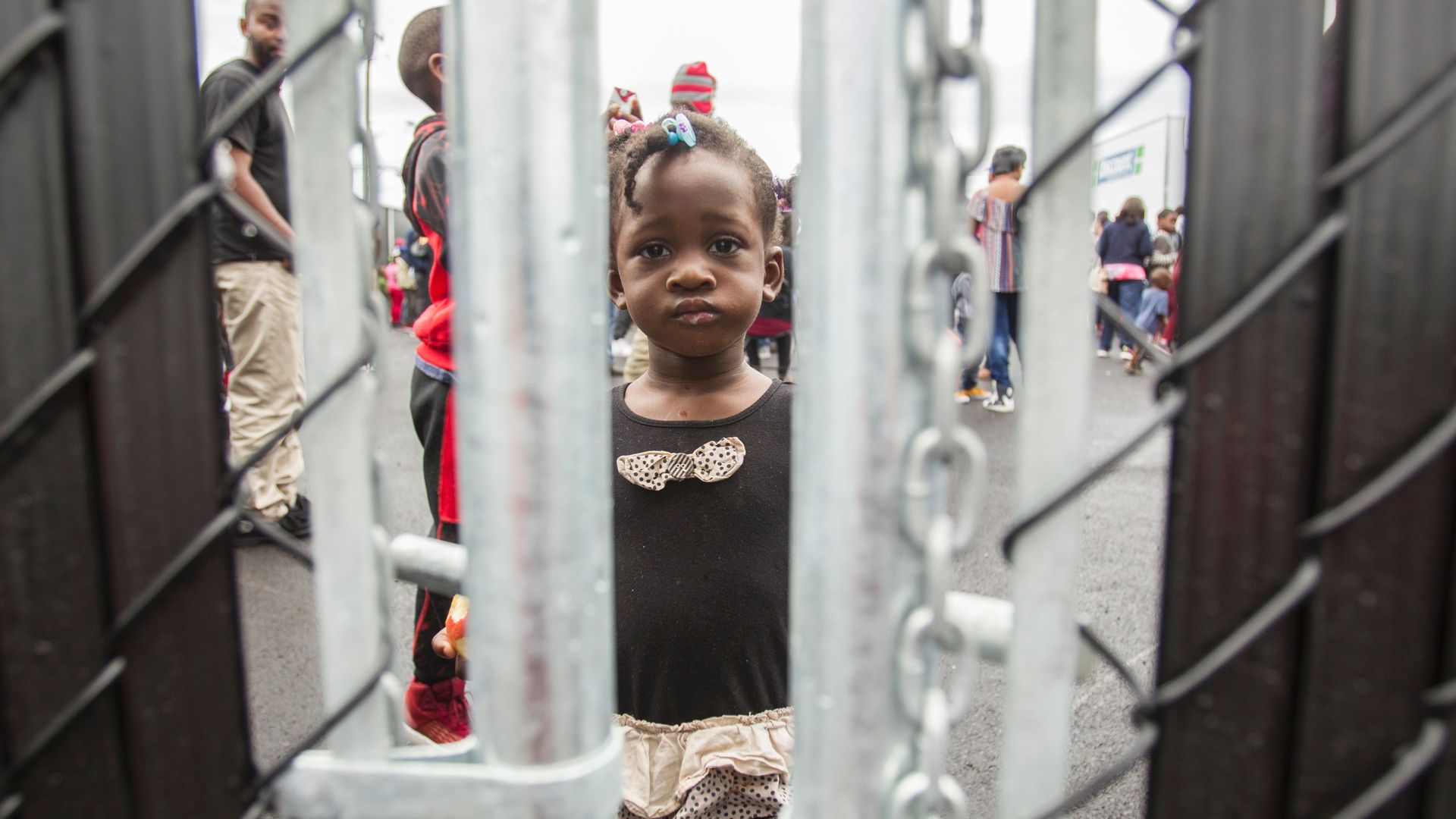 Little black girl with pink bows looks through chain linked fence with a frown. 