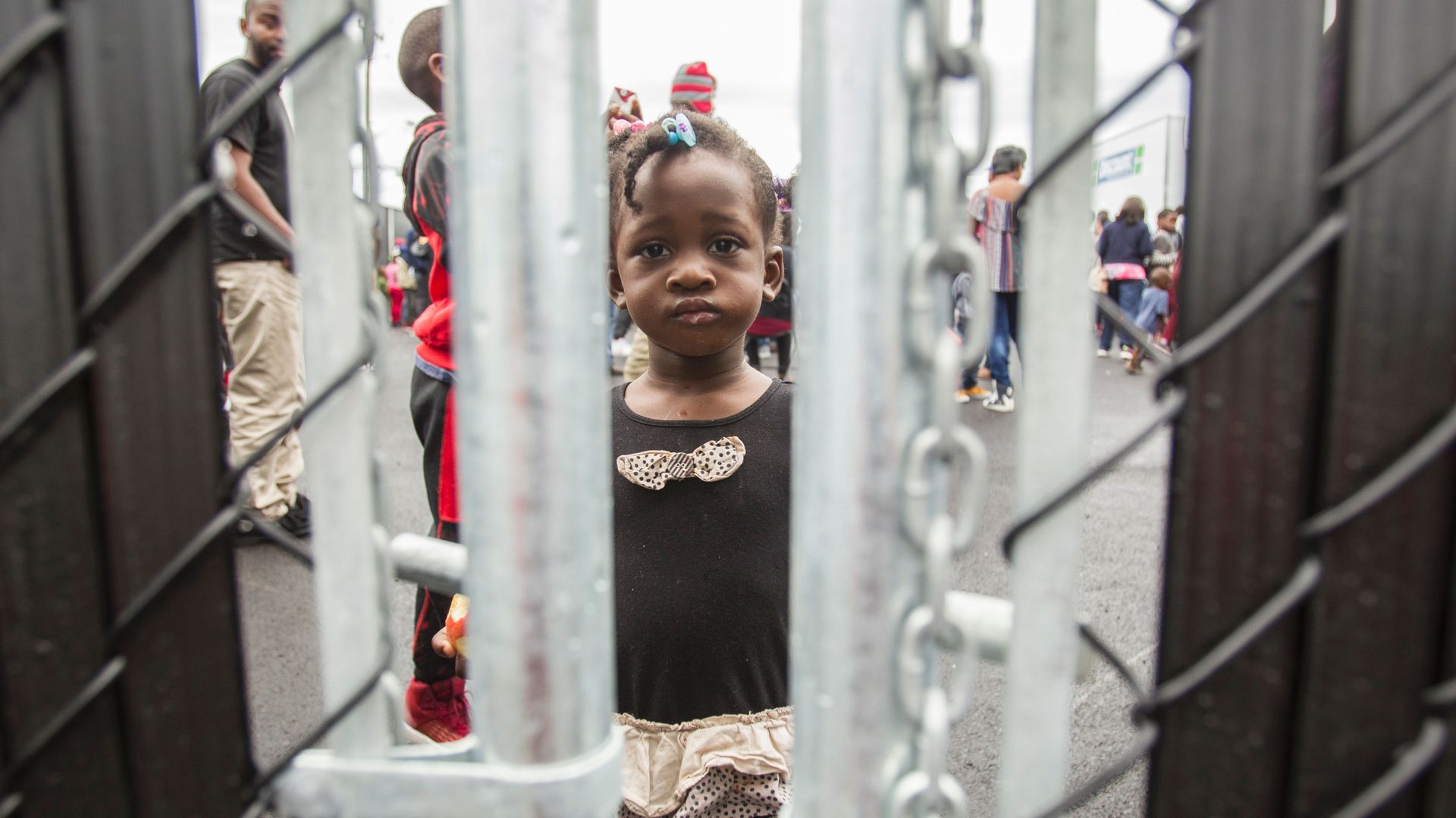 Little black girl with pink bows looks through chain linked fence with a frown.