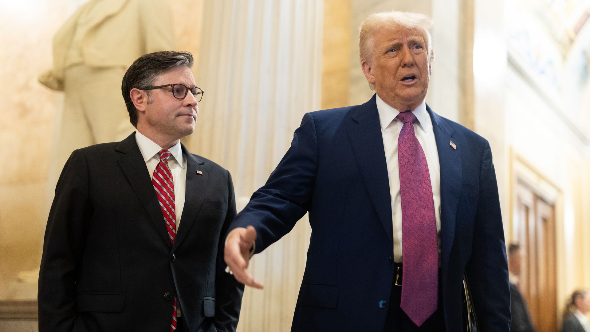 House Speaker Mike Johnson wearing a black suit and Trump wearing a blue suit standing in a white marble hallway.