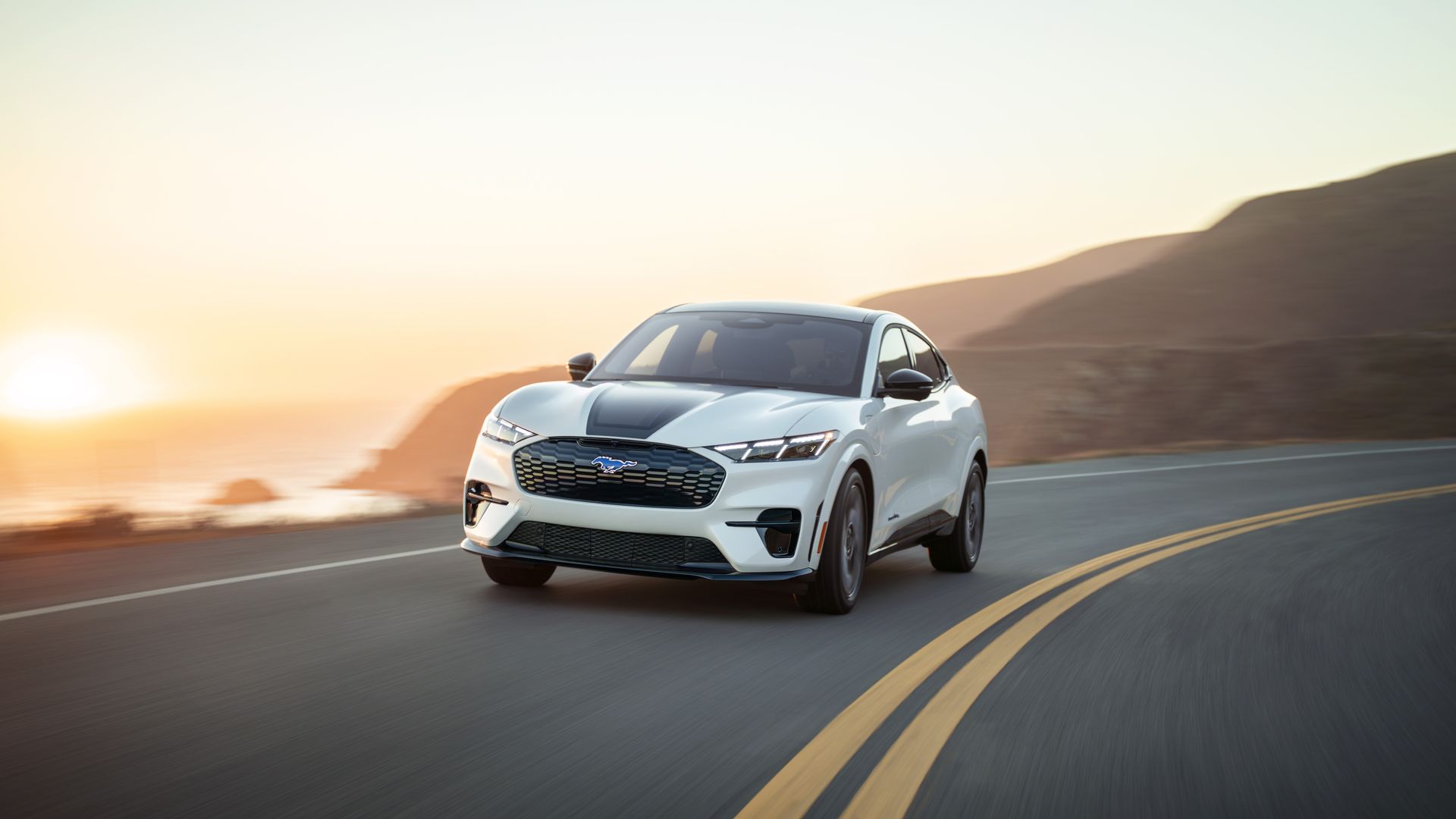 A version of a white Ford Mustang Mach-E driving on a winding road at sunset with hills and ocean in the background.