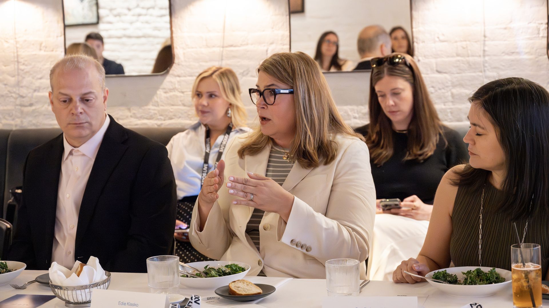 Business meeting in a bright restaurant: a woman with glasses speaks animatedly at a table with salads, bread, and water, others listening; white brick walls and modern mirrors in the background.