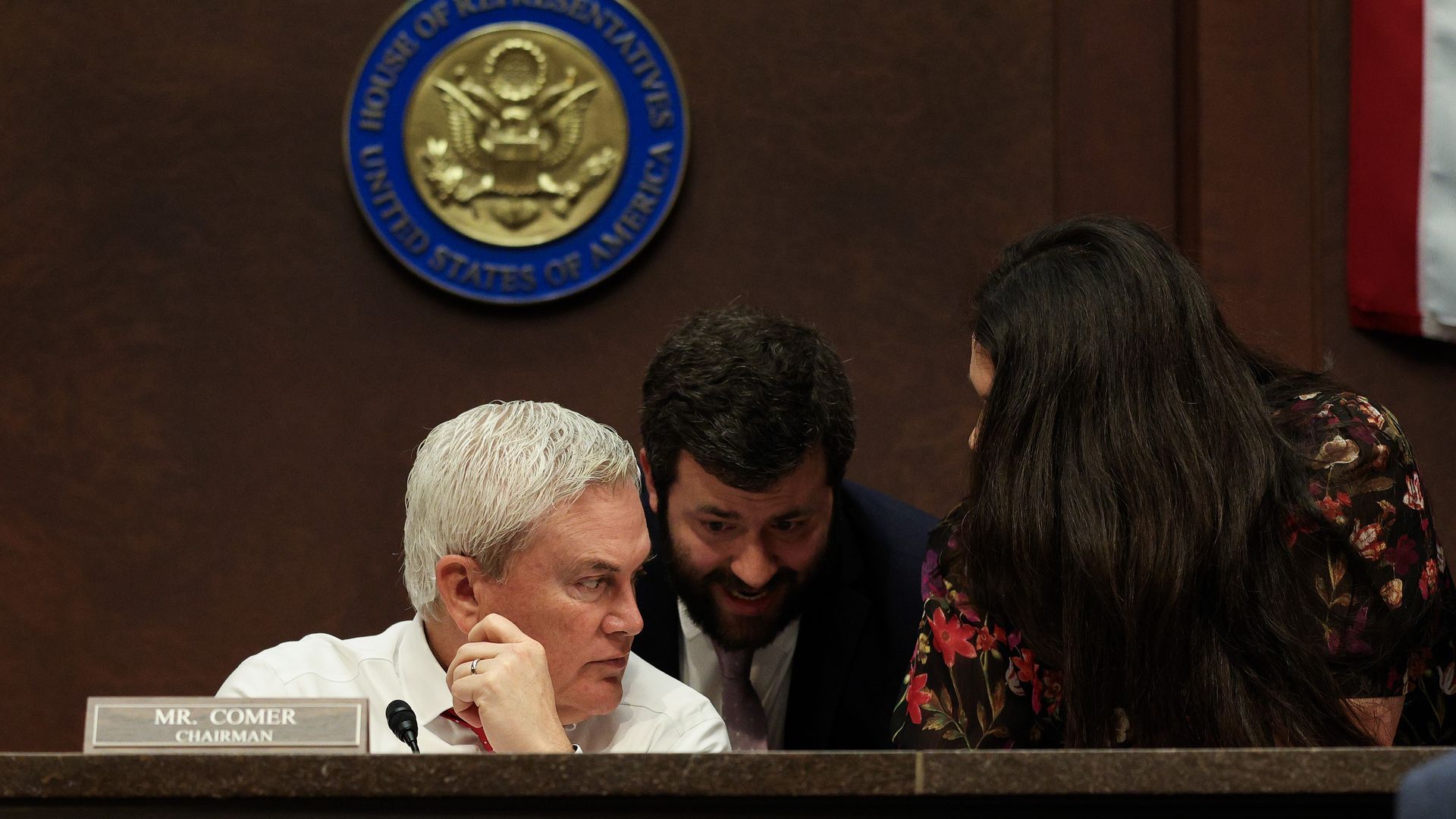 Three people conversing closely behind a desk with a sign reading "Mr. Comer Chairman" and a House of Representatives seal on a wood-paneled wall, one in a white shirt, one in a dark suit, one in floral top.
