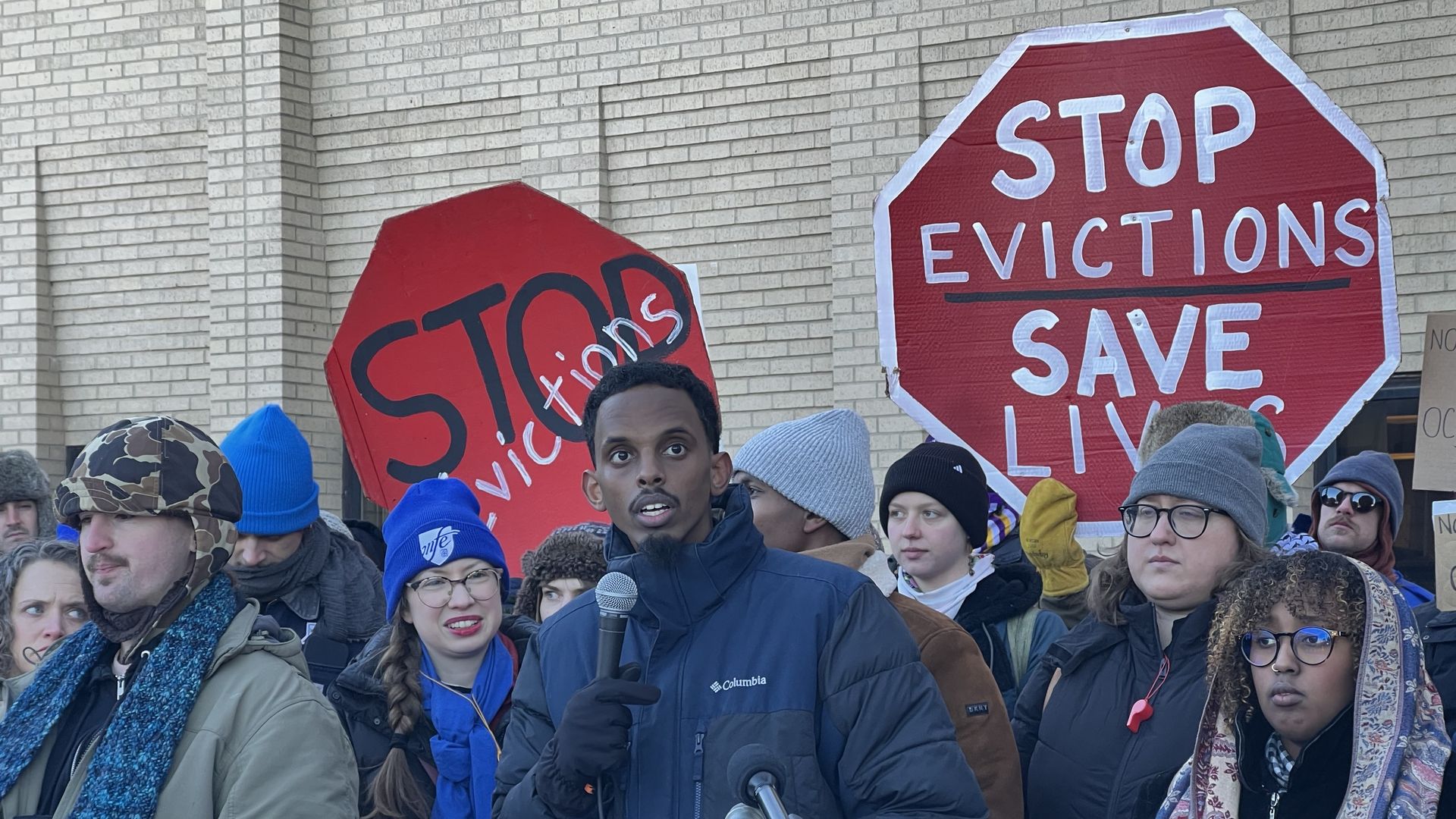 A diverse group of people bundled in winter clothes at a rally protesting evictions, holding red stop-sign-shaped signs that say "STOP evictions" and "STOP EVICTIONS SAVE LIVES".