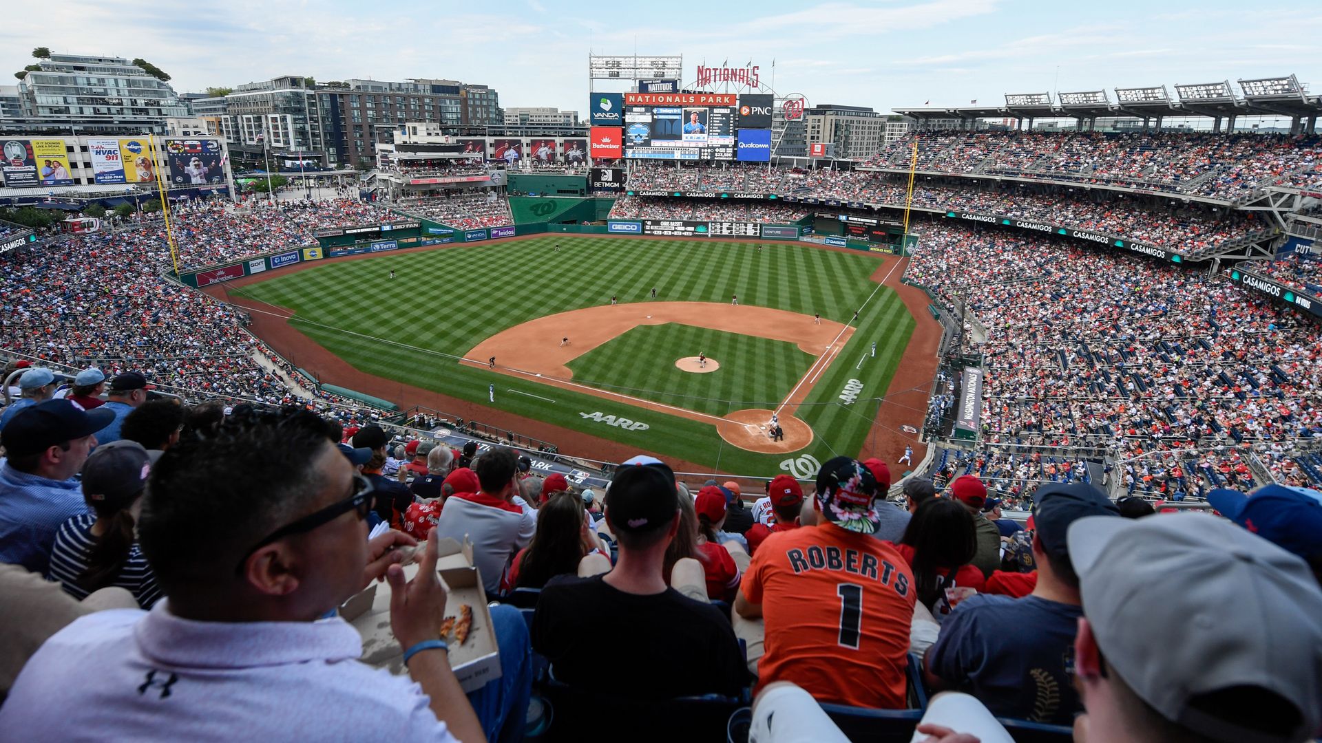 Wide view of a baseball game at Nationals Park: packed stands with red-clad fans, green field, players on the diamond, and a large scoreboard against a clear blue sky.