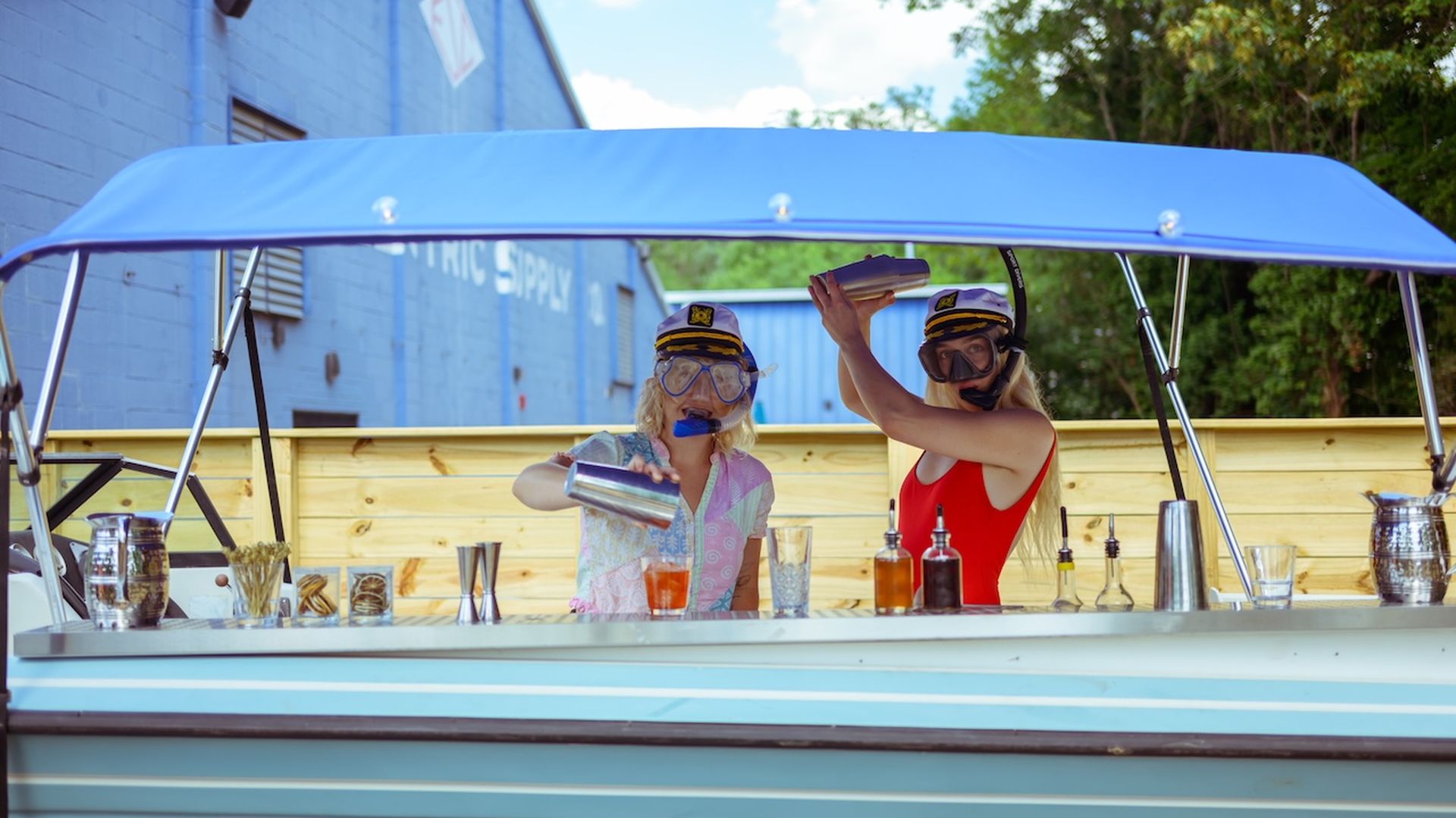 Two women wearing captain hats and snorkeling goggles mix cocktails on a retro bar boat under a blue canopy, pouring from shakers into glasses beside bottles and bar tools.