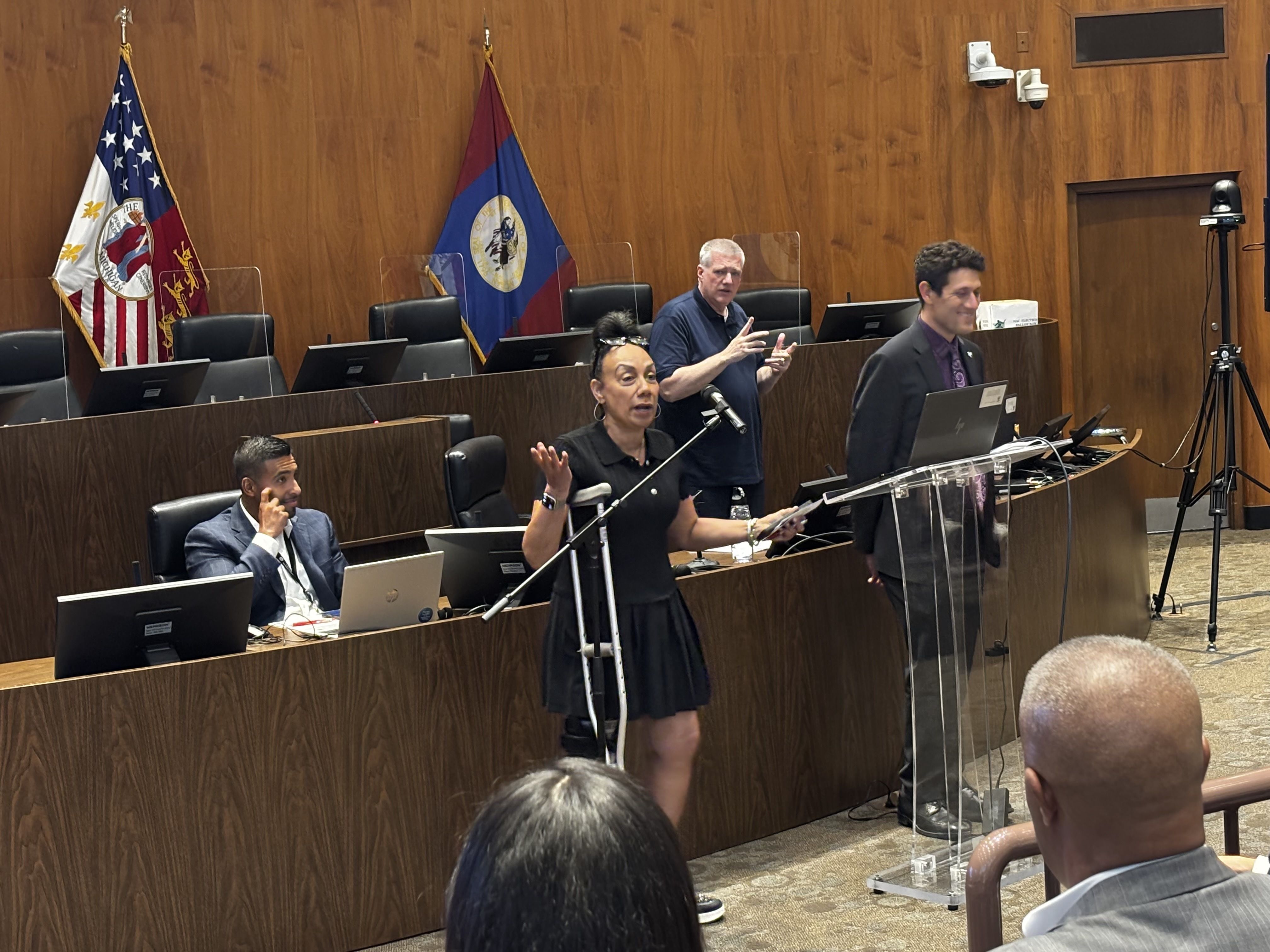 Wood-paneled council chamber with flags behind. Officials at desks with laptops and microphones, acrylic barriers. A woman speaks; a man gestures; a suited man smiles. A camera on a tripod to the right.