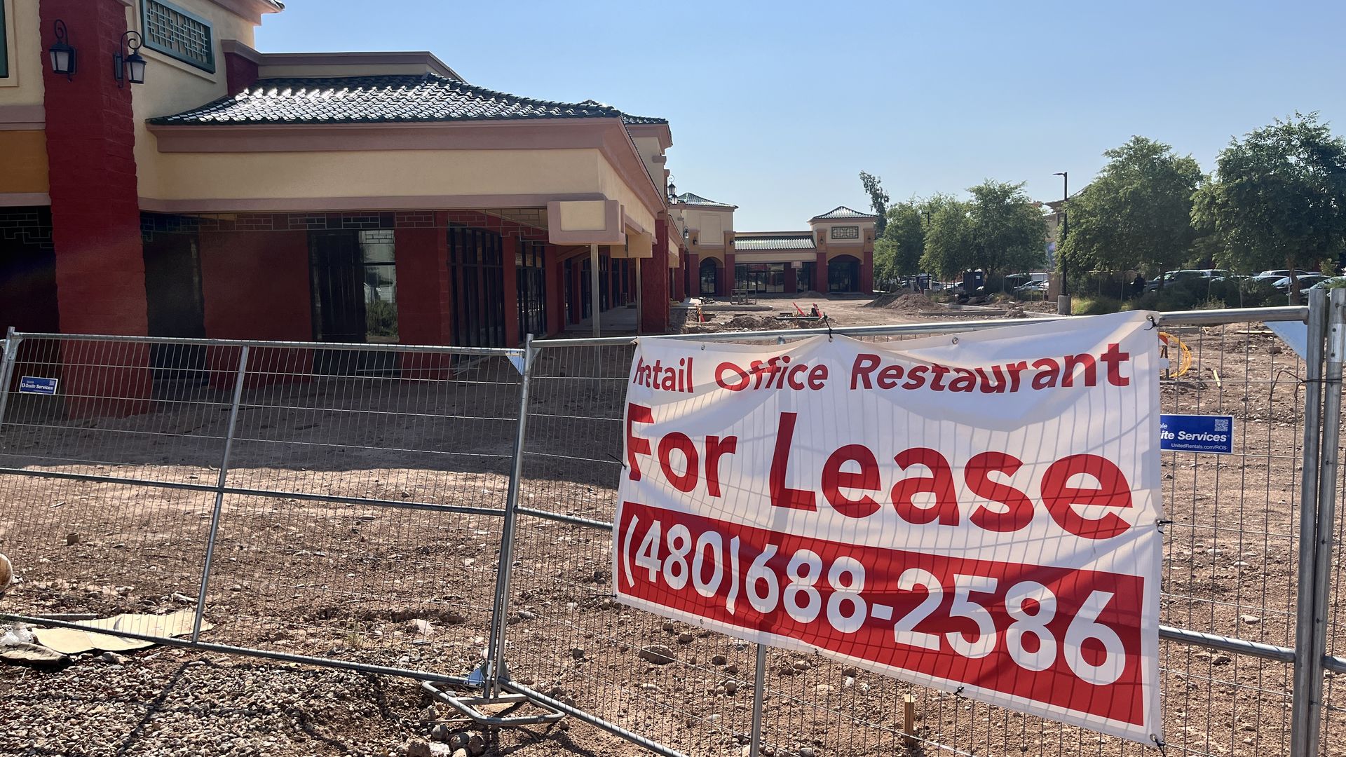 A sign that says, "For lease" with a strip mall that has a decorative roof int he background. 
