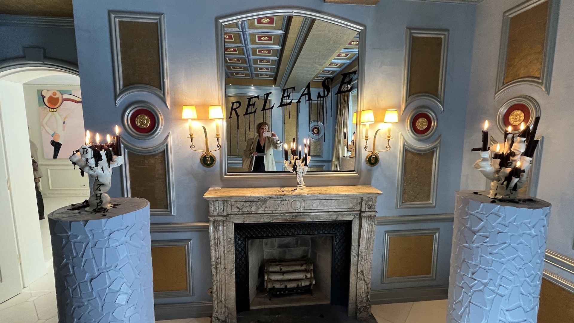 Blue paneled room with a marble fireplace and mirror above it. The word "RELEASE" appears in the reflection. A person in a beige coat takes a selfie, flanked by two lit pedestals.