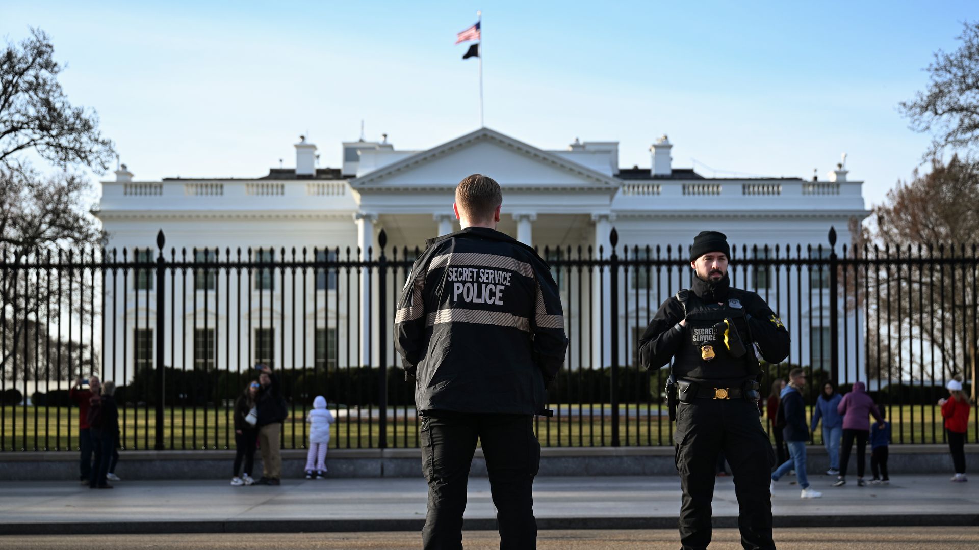 Two Secret Service officers stand with their backs to the camera in black uniforms, in front of the White House behind a tall iron fence, with an American flag and visitors nearby.