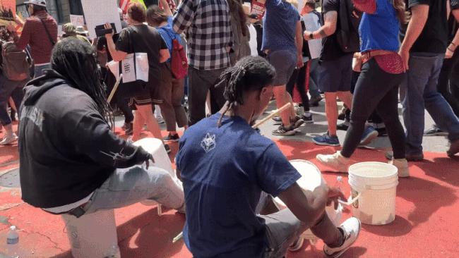 Photo of two people playing buckets like drums while people march by. 