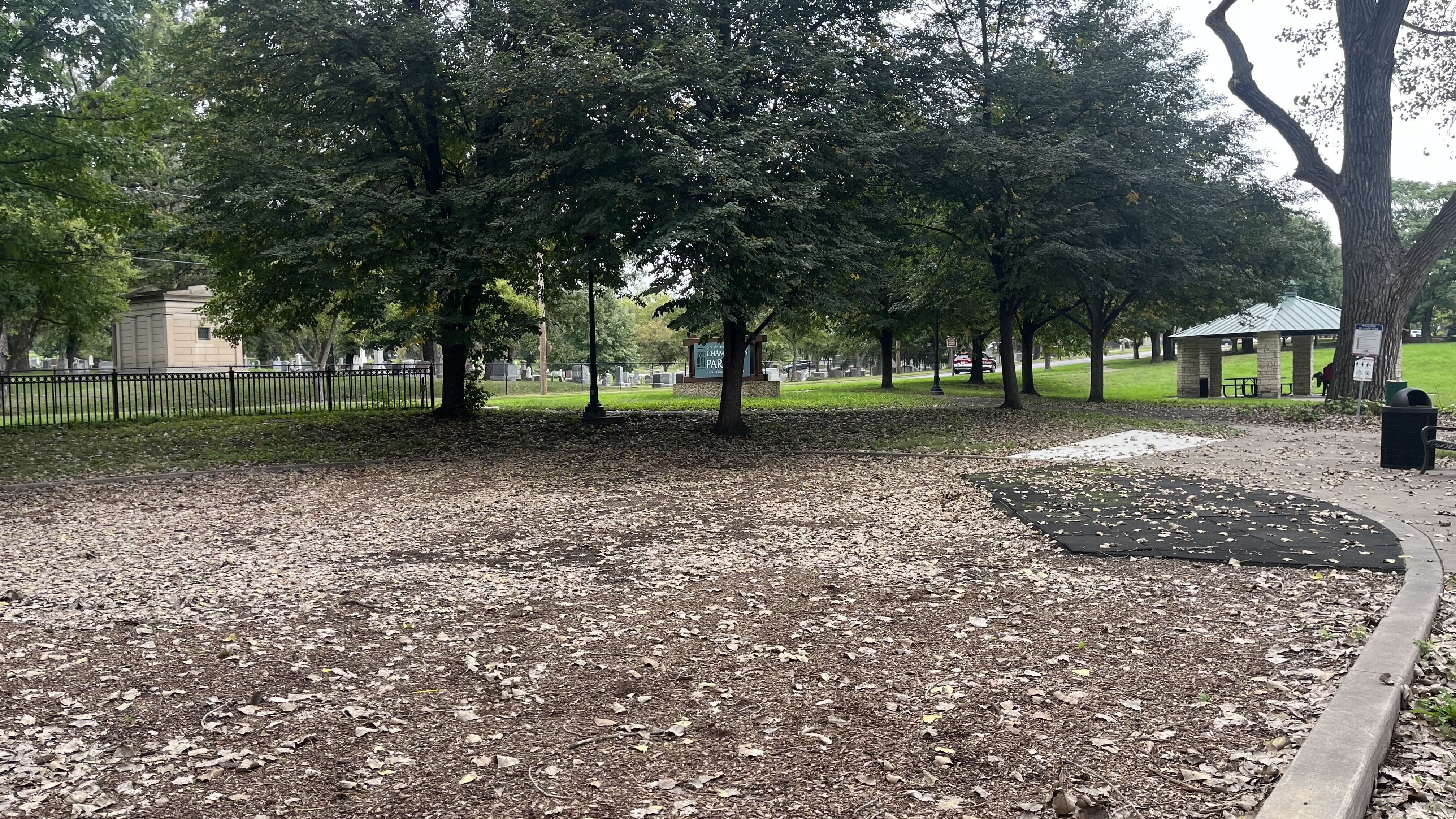 Park scene with dense trees and fallen leaves, a curved path on the right, a beige mausoleum on the left, a black fence, and a small pavilion with benches on the right.