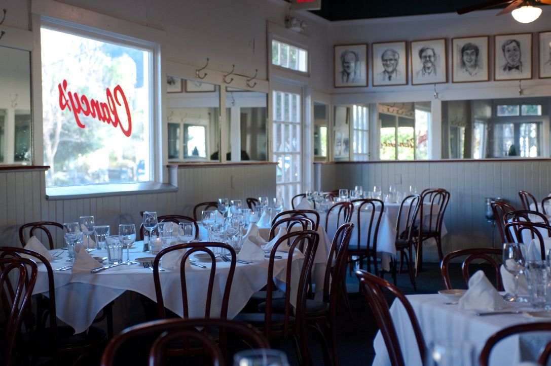 An empty dining room with tables covered in flatware, napkins, glasses and white tablecloths.