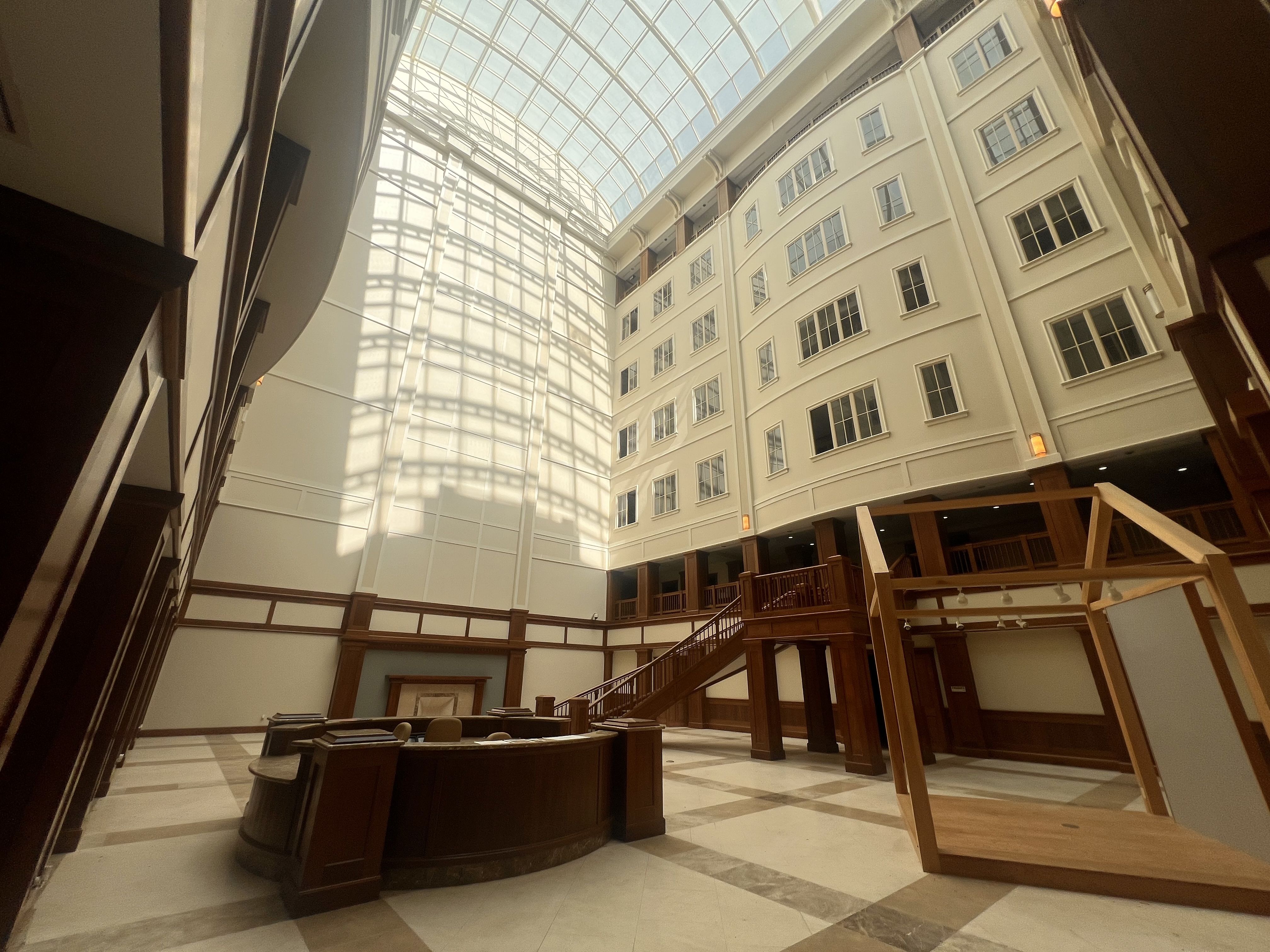Indoor atrium with cream walls, tall curved windows, and a glass skylight overhead. A round dark wood reception desk sits on the marble floor; wooden railings and stairs surround the open space.