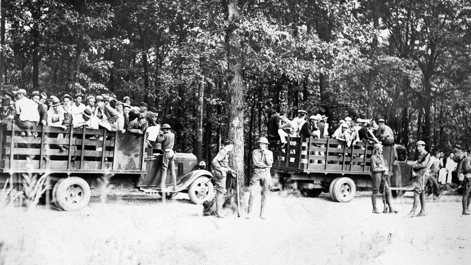 A black and white photo of striking workers standing in the beds of trucks as they arrive at a prison camp in the woods