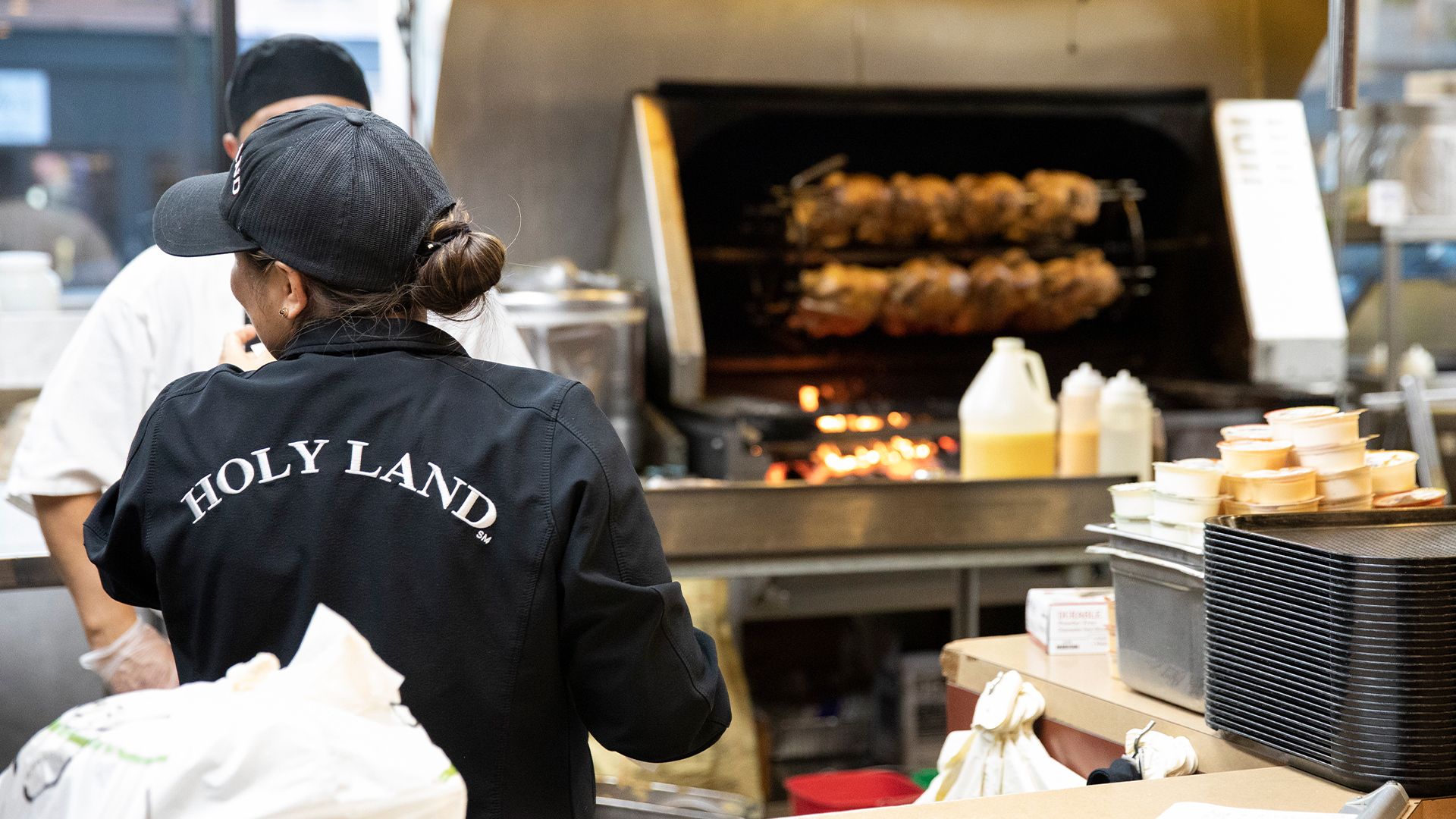 An employee wearing "Holy Land" branded clothing working near a grill area, where rotisserie chickens cook in the background.