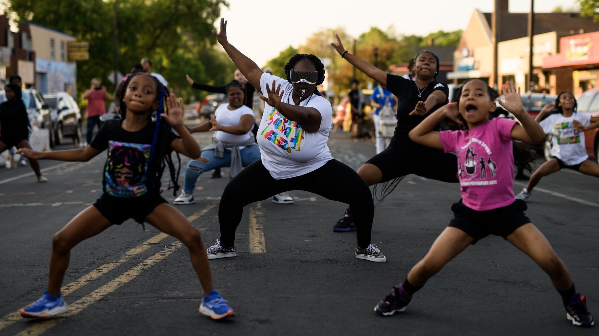 A group of dancers, two young girls in the front, and adults in the back, perform a dance in a street during a parade