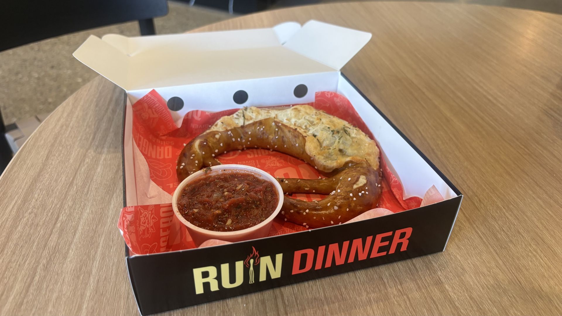 A large pretzel in a to-go box next to a side or marinera.