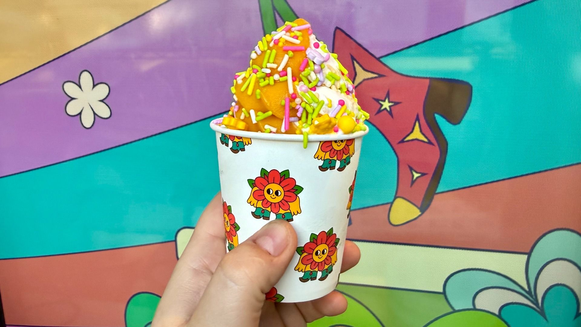 Hand holding a white cup printed with sunflowers and cartoon characters, topped with orange soft serve and multicolored sprinkles. A vibrant mural with purple, teal, and orange fills the background.