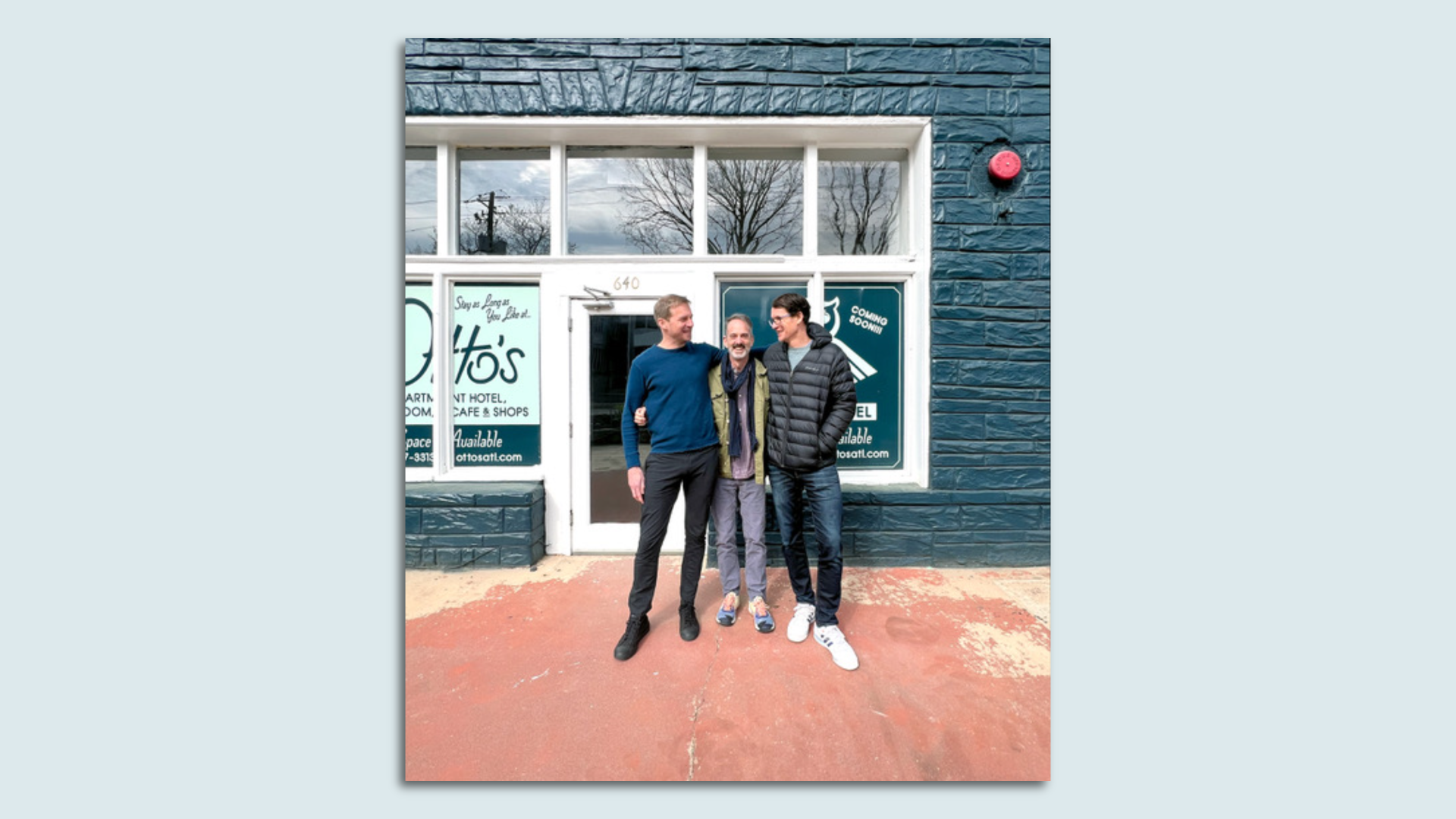 Three smiling men with their arms around each other stand outside a dark green vacant storefront 