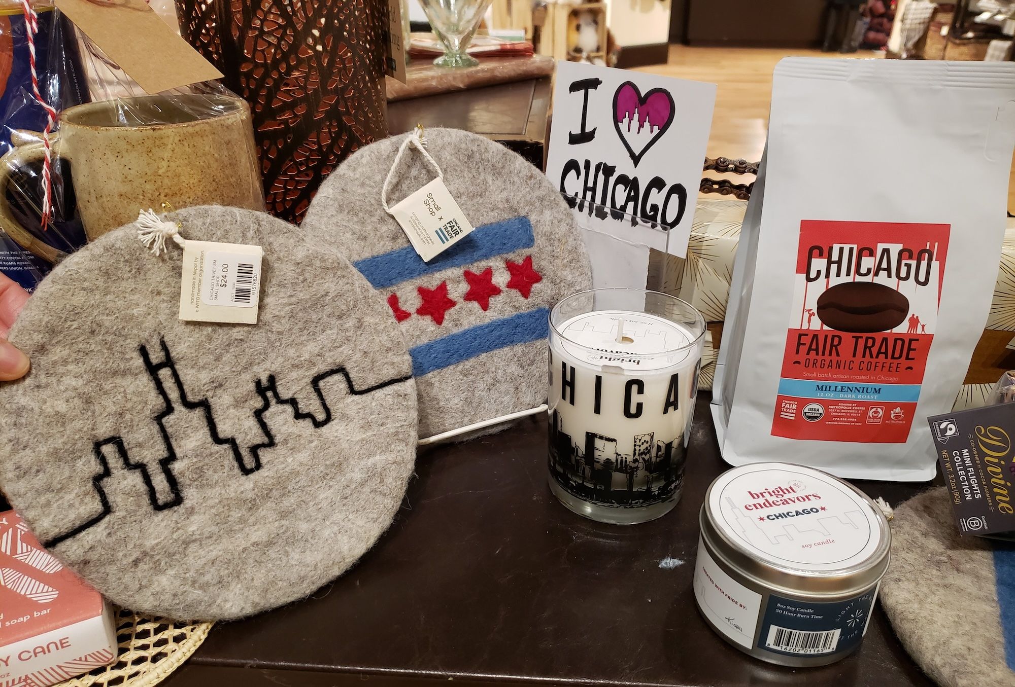 Display of Chicago-themed items including felt coasters with skyline and flag designs, a candle with Chicago skyline, a bag of Chicago Fair Trade coffee, and a sign saying "I ♥ Chicago".