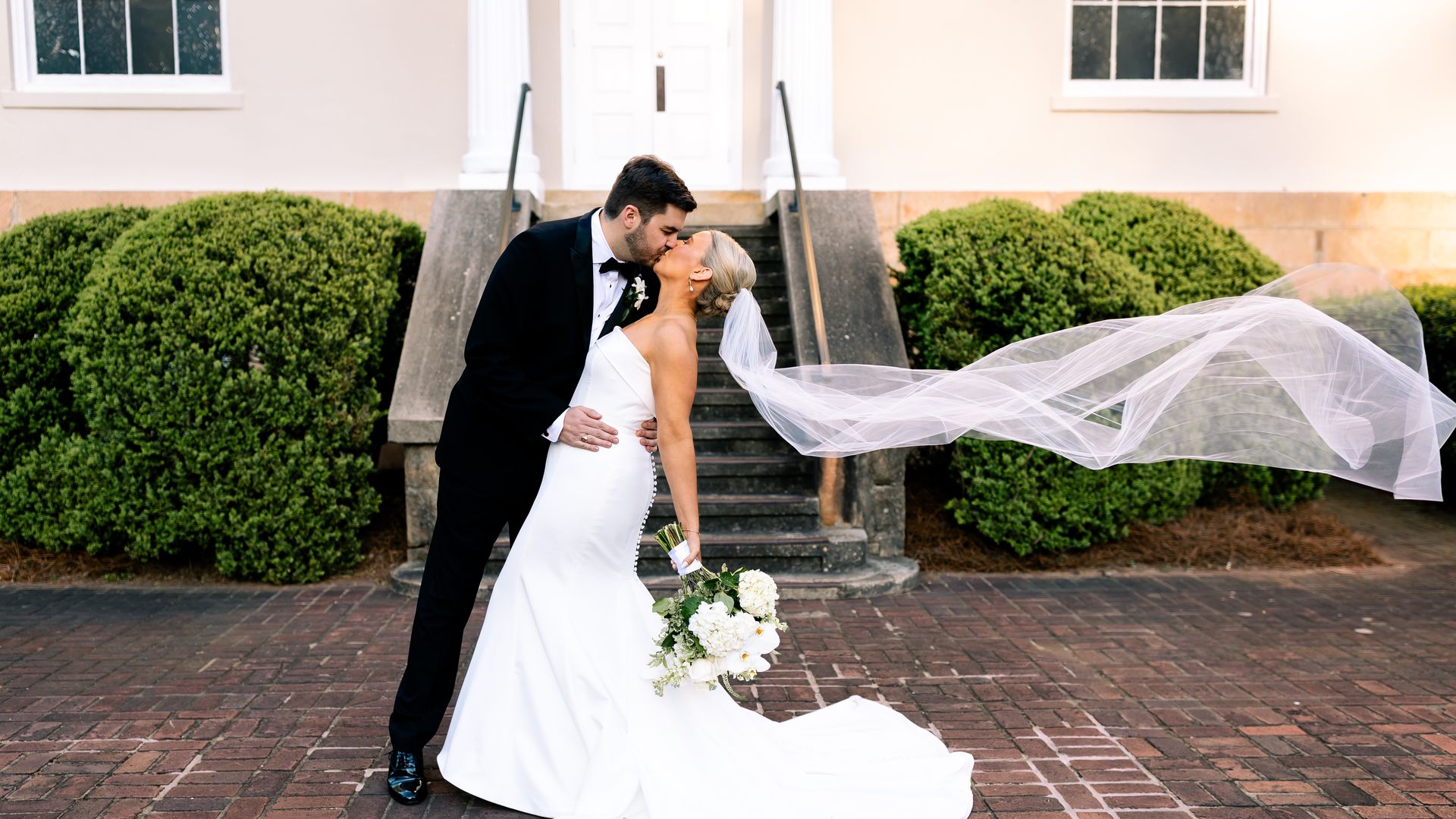 Bride in white gown with long veil and bouquet kisses groom in black tuxedo outdoors on brick path with green bushes and steps behind them.