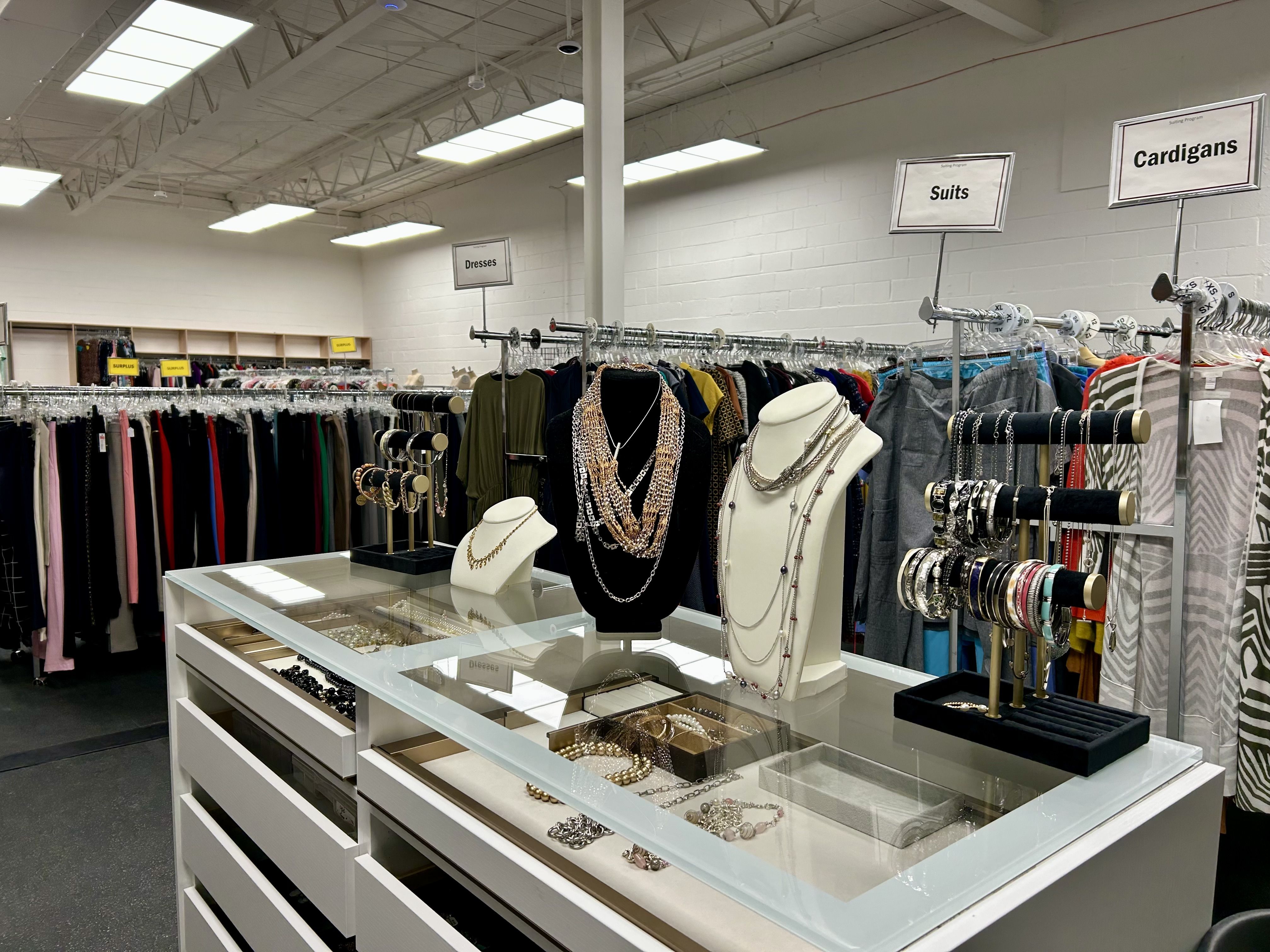 Jewelry display with necklaces and bracelets on stands in front of racks labeled Dresses, Suits, and Cardigans in a well-lit clothing store.