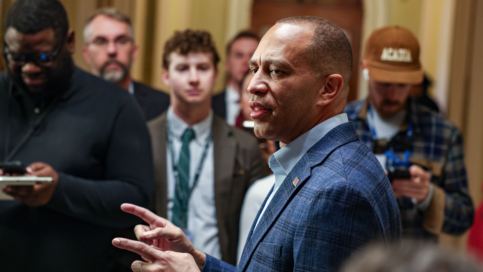 House Minority Leader Hakeem Jeffries, wearing a blue suit and speaking to reporters in a beige hallway.