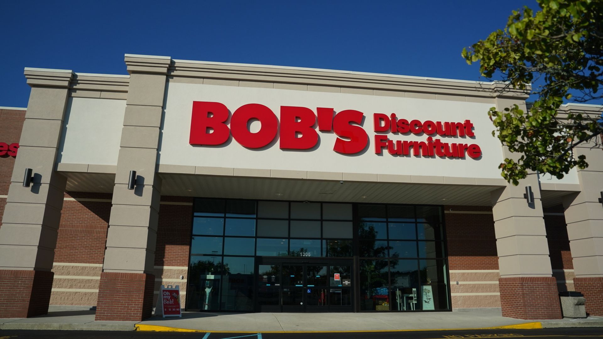 Front entrance of Bob's Discount Furniture store with large red sign on beige wall under clear blue sky, trees partially visible on right.