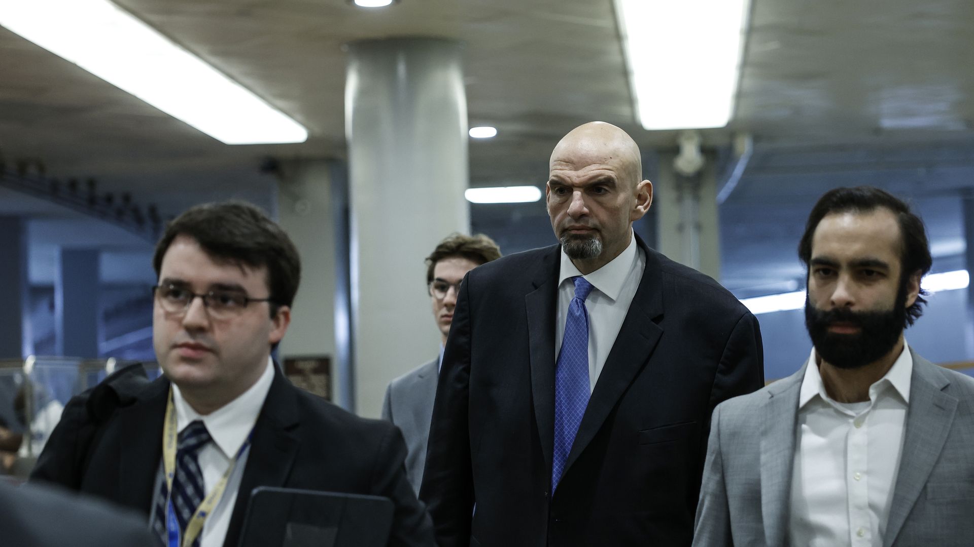 Sen. John Fetterman, wearing a dark gray suit, white shirt and blue tie, walks through the Senate basement with staffers.