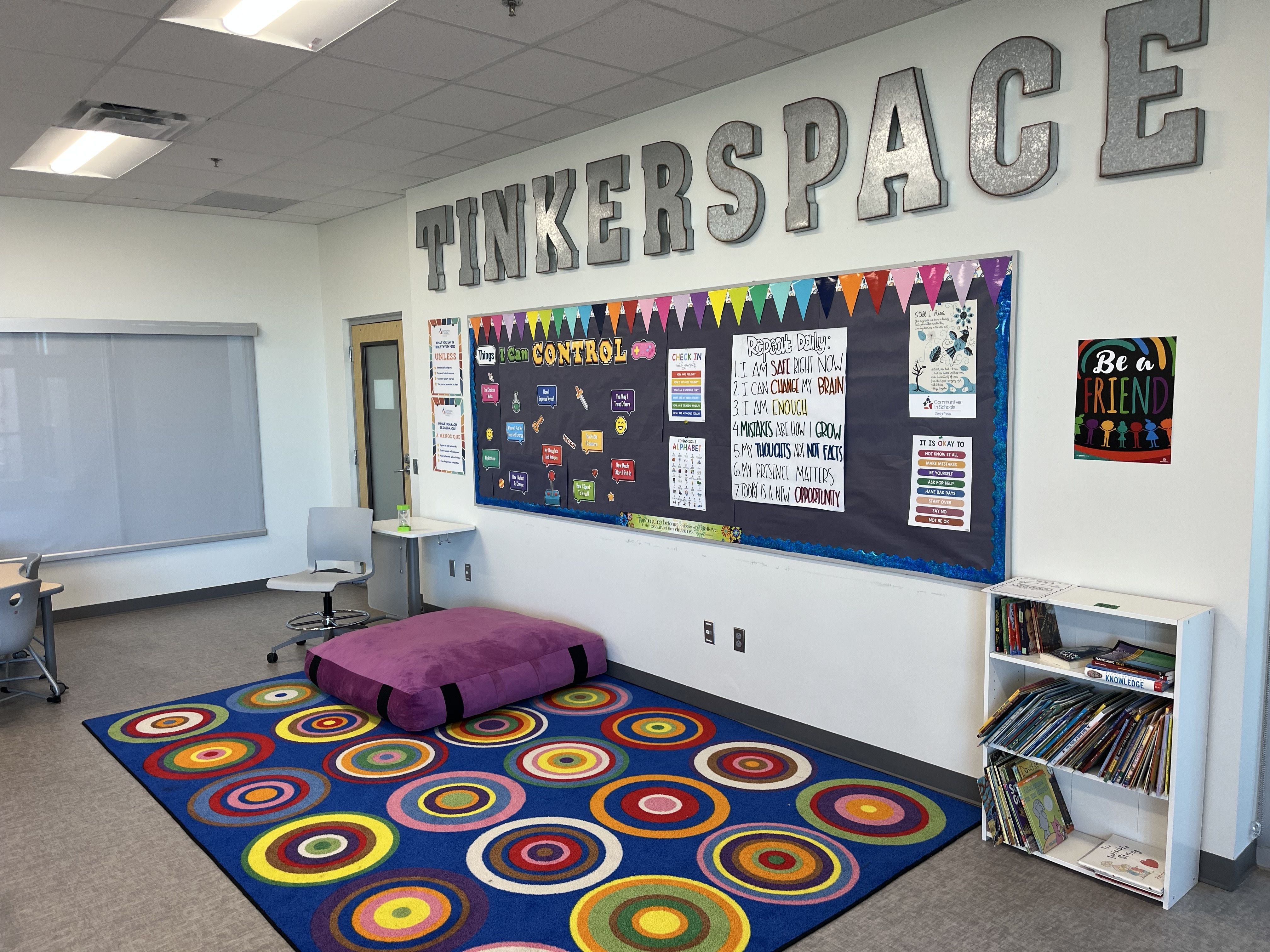 Colorful classroom corner with a blue rug featuring multicolored circles, purple cushion, white chair, bookshelf with books, black bulletin board with motivational posters, and large metallic letters spelling TINKERSPACE.