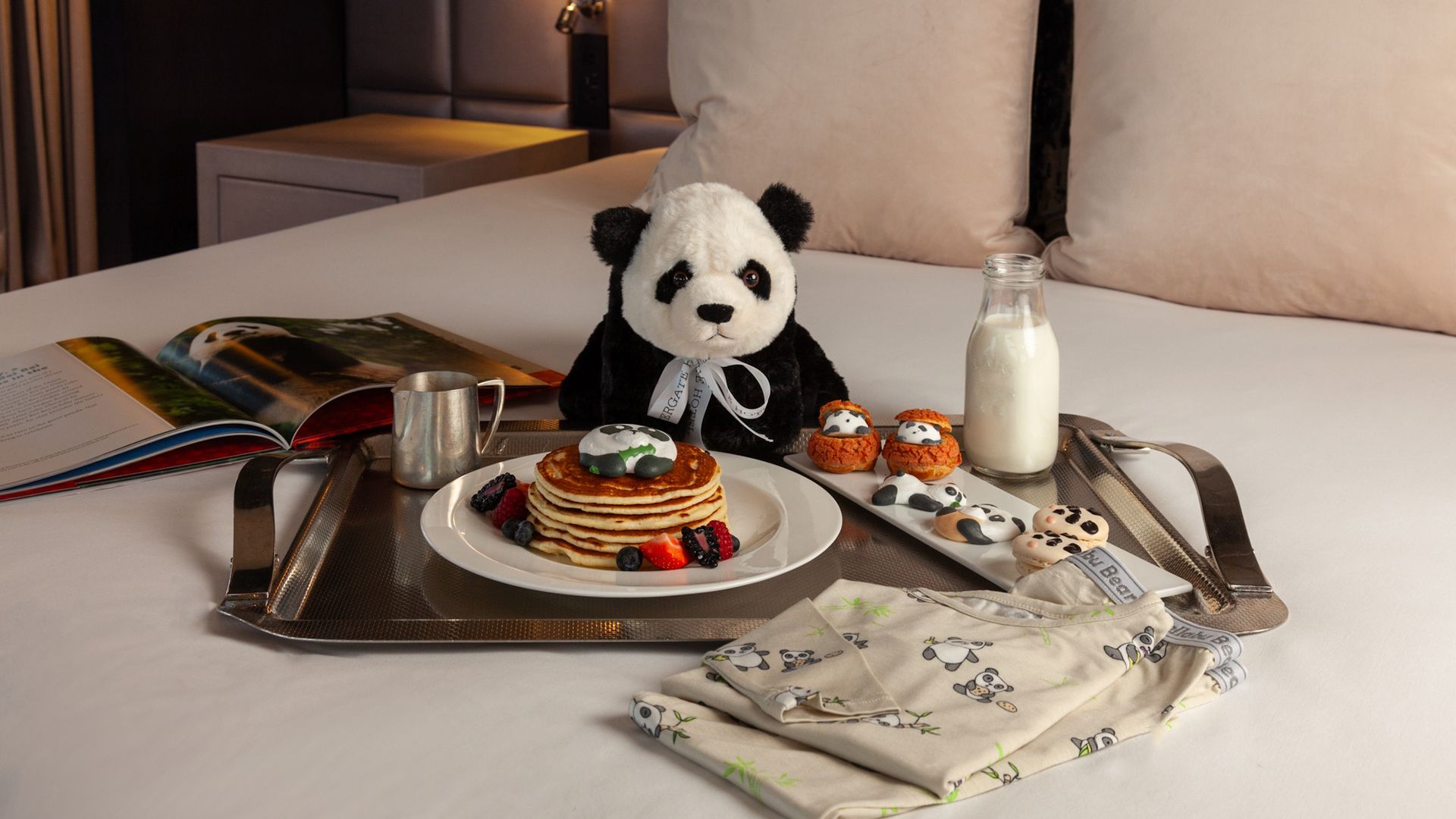 A panda themed hotel package on a bed with a panda stuffed animal, panda pajamas and panda sweets.