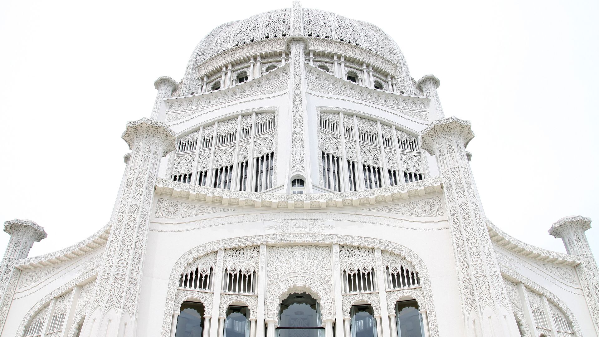 White, intricately carved building with arabesque patterns, tall arched windows, and a large central dome; ornate stonework dominates the facade against a pale sky.
