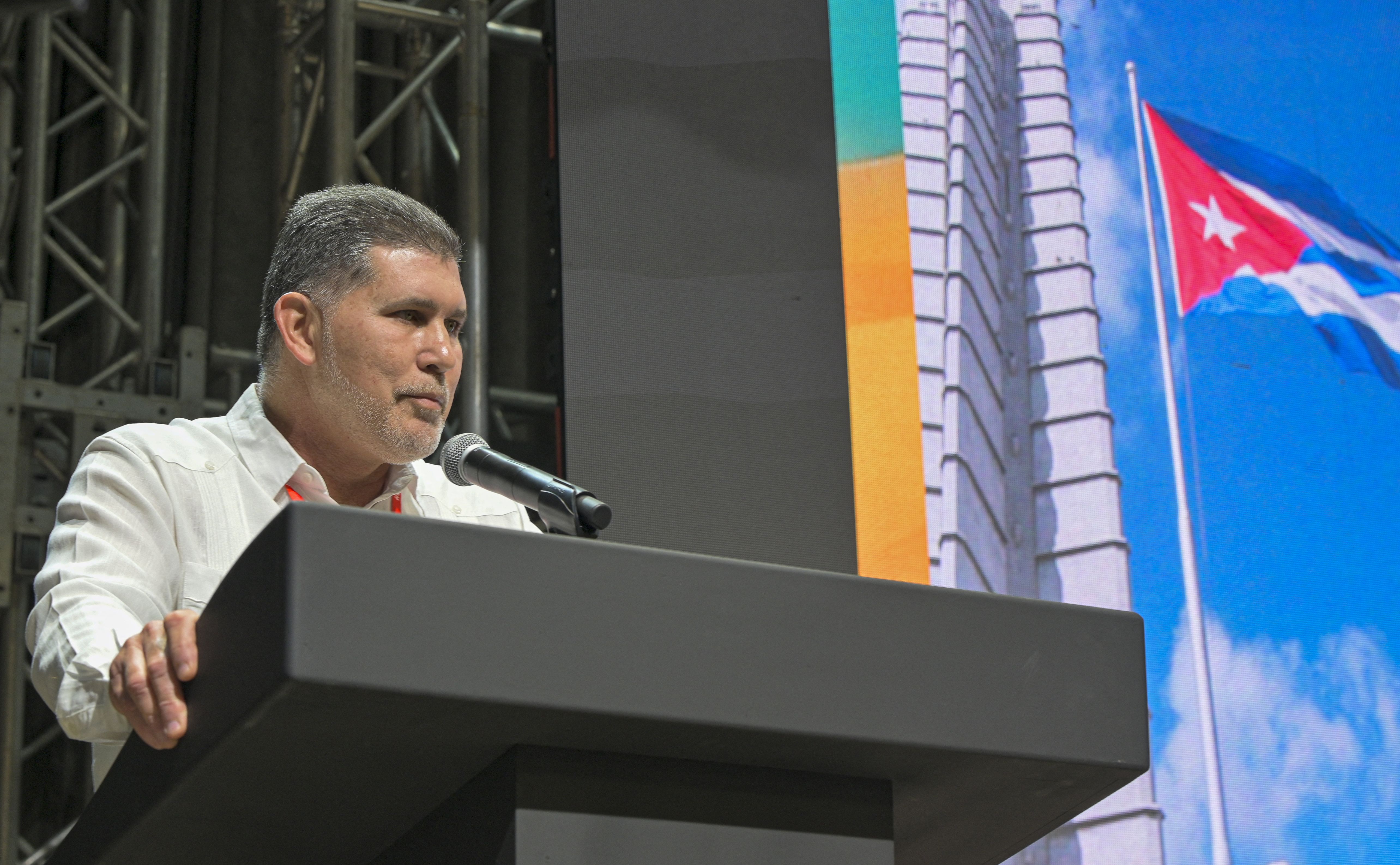Man in a white shirt speaks at a black podium with a microphone on a stage. Behind him, a large screen shows a building and a waving Cuban flag against a blue sky.