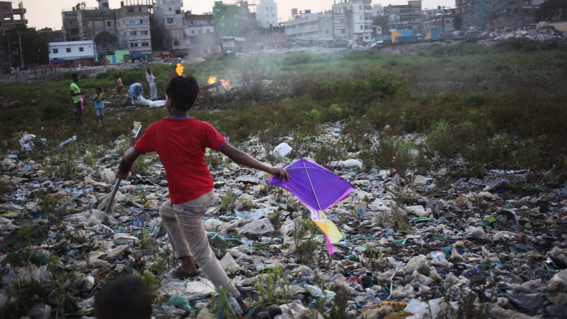 In this image, a boy in a bright shirt carries a kite while running over a field of garbage.