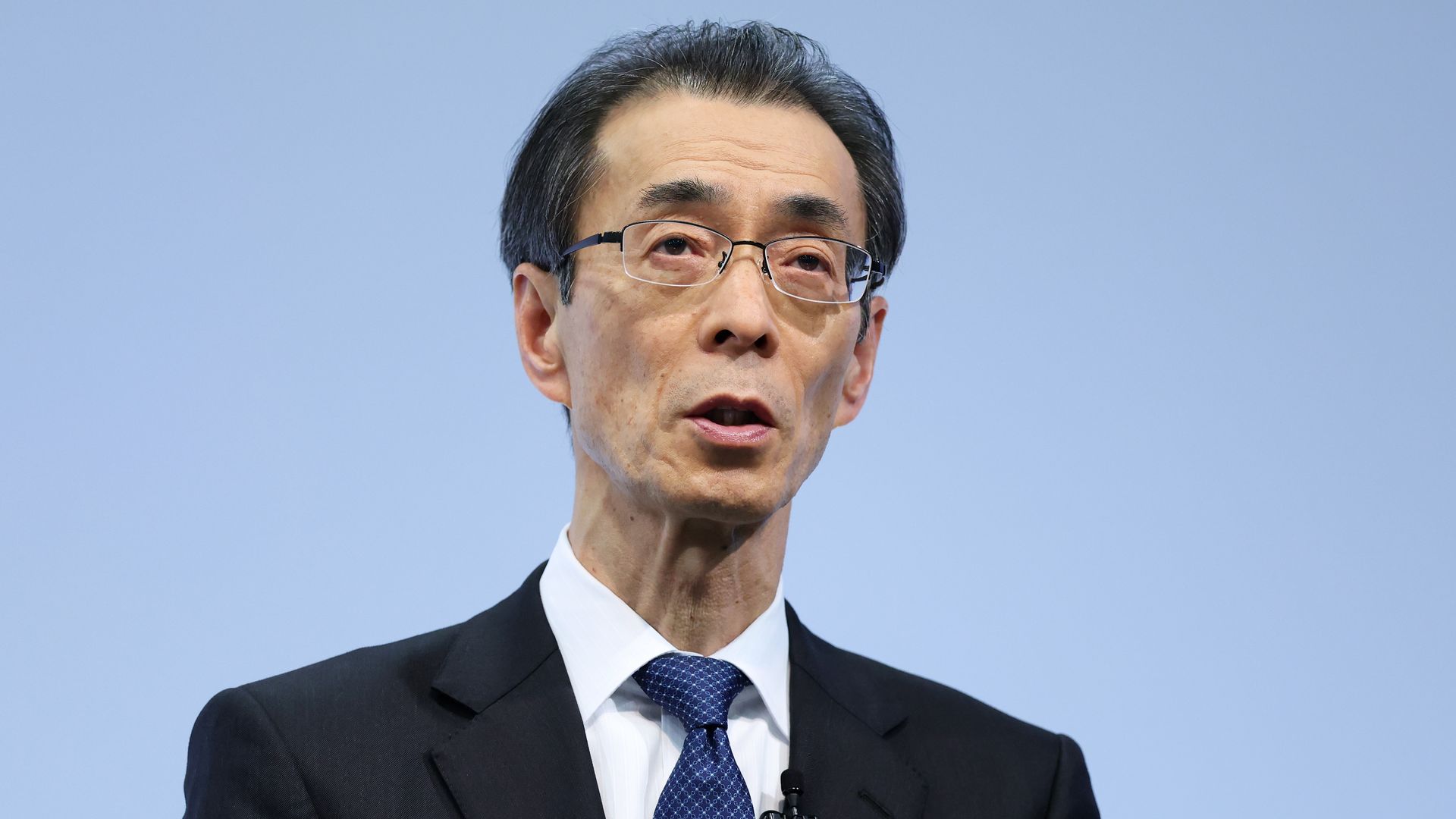 Close-up of a person in a dark suit, white shirt, and blue patterned tie, wearing glasses with a lapel microphone, speaking against a clear blue sky.