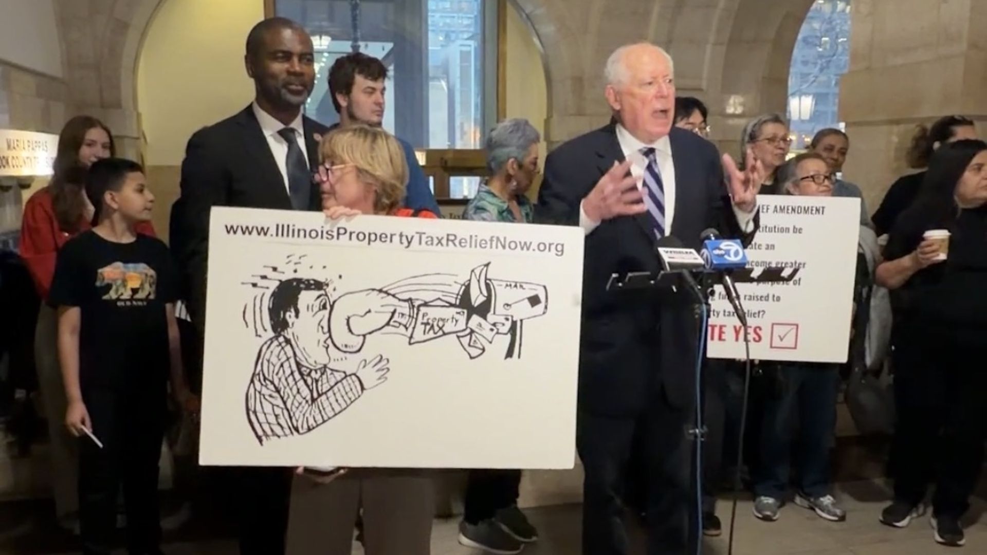 Group stands in a marble lobby; a man and woman hold a sign reading www.IllinoisPropertyTaxReliefNow.org with a cartoon of a fist punching a tax figure; a suited man speaks at a microphone.