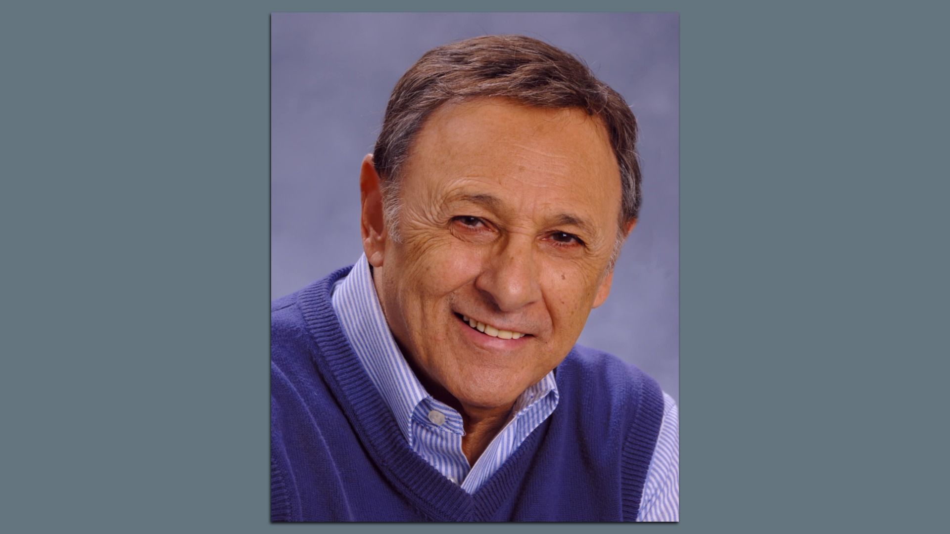 Portrait of a smiling older man with short brown hair, wearing a blue sweater vest over a light blue striped shirt, against a plain gray background.