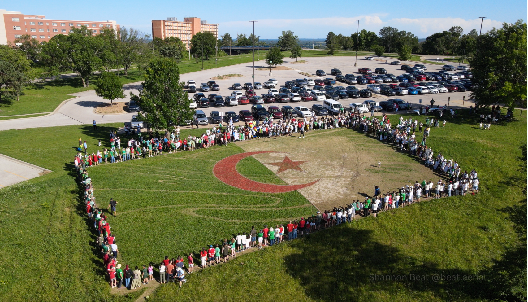 Aerial view of a large crowd forming a curved line around a red crescent and star on a grassy field, with a parking lot full of cars and buildings in the distance.