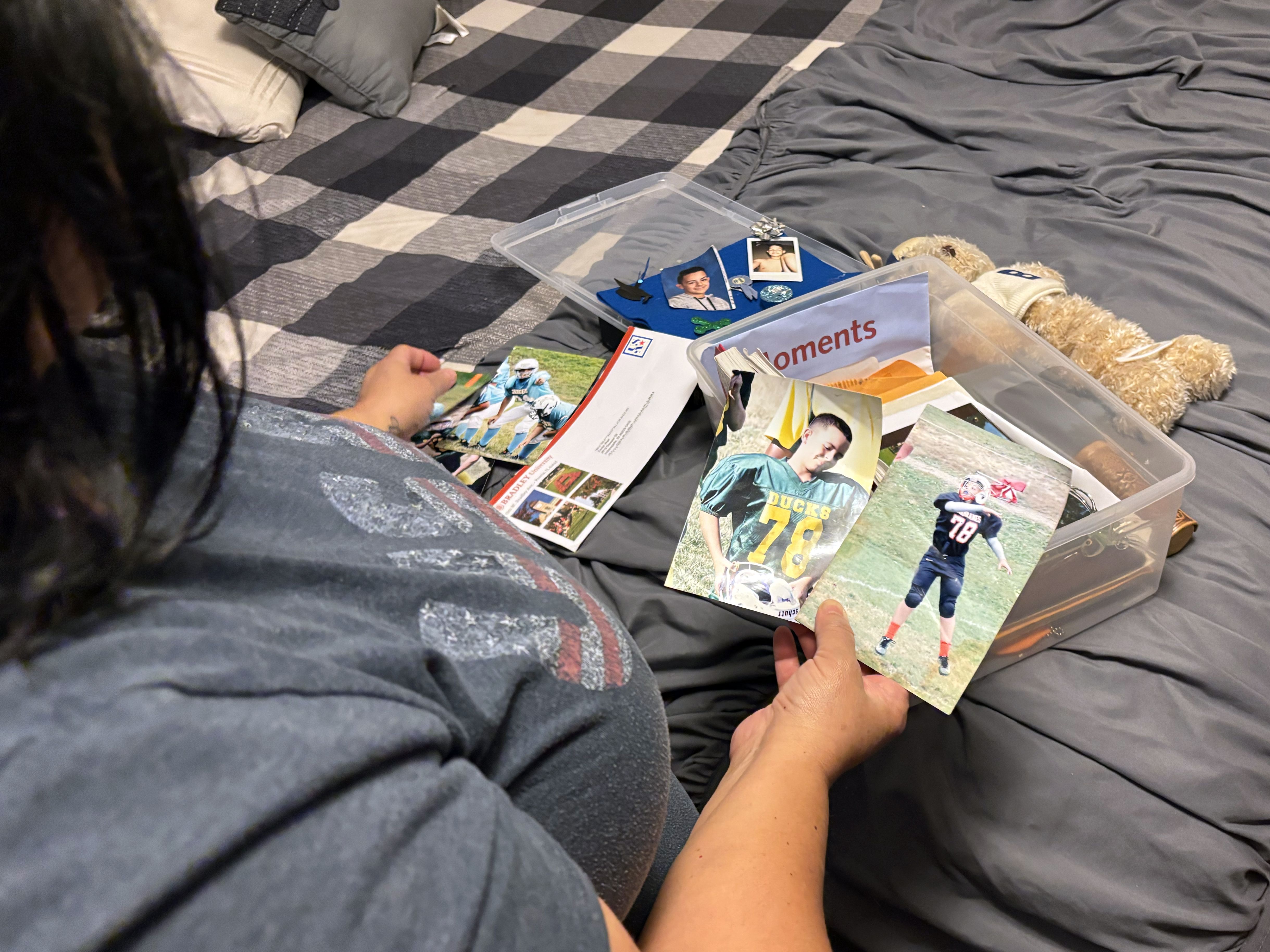 Person on a bed looking through photos and memorabilia in a clear plastic box labeled "Moments," including football pictures, keys, and a teddy bear on a gray and black checkered bedspread.