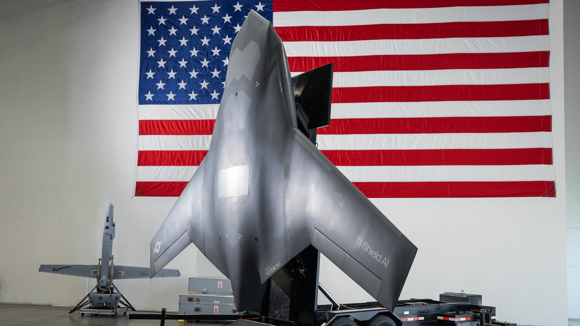 Gray drone with Shield AI and U.S. Air Force logos on a trailer inside a hangar, large American flag hanging behind it.
